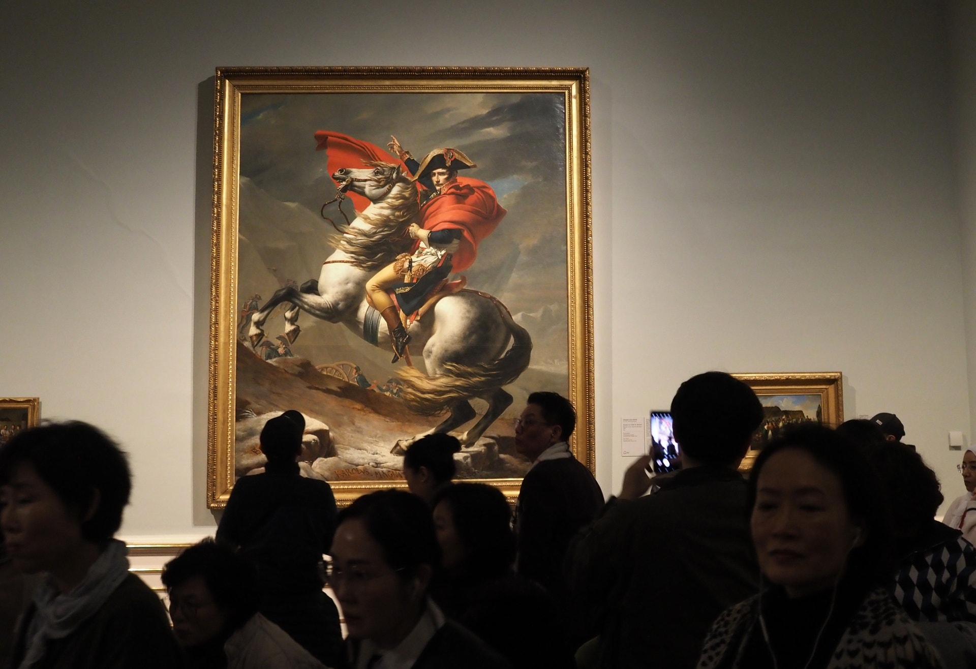 Ett klassiskt måleri föreställande en soldat på en vit häst med röd kappa. Målningen hänger på en vägg i ett museum där flera besökare betraktar verket.
