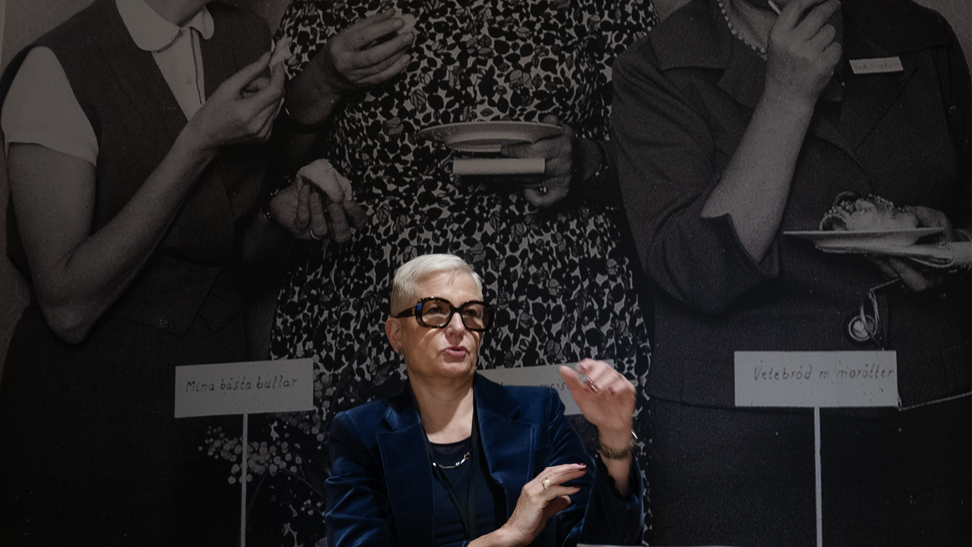 Korthårig blond kvinna med stora, mörka glasögon, Nina Jönsson, koncernchef för Ica, sitter framför en fotovägg och gestikulerar med händerna.