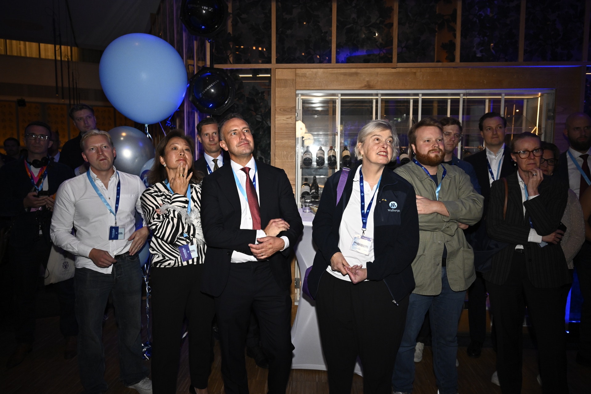 En grupp människor står samlade inomhus på ett evenemang och tittar uppåt mot något, omgivna av ballonger och festlig belysning.