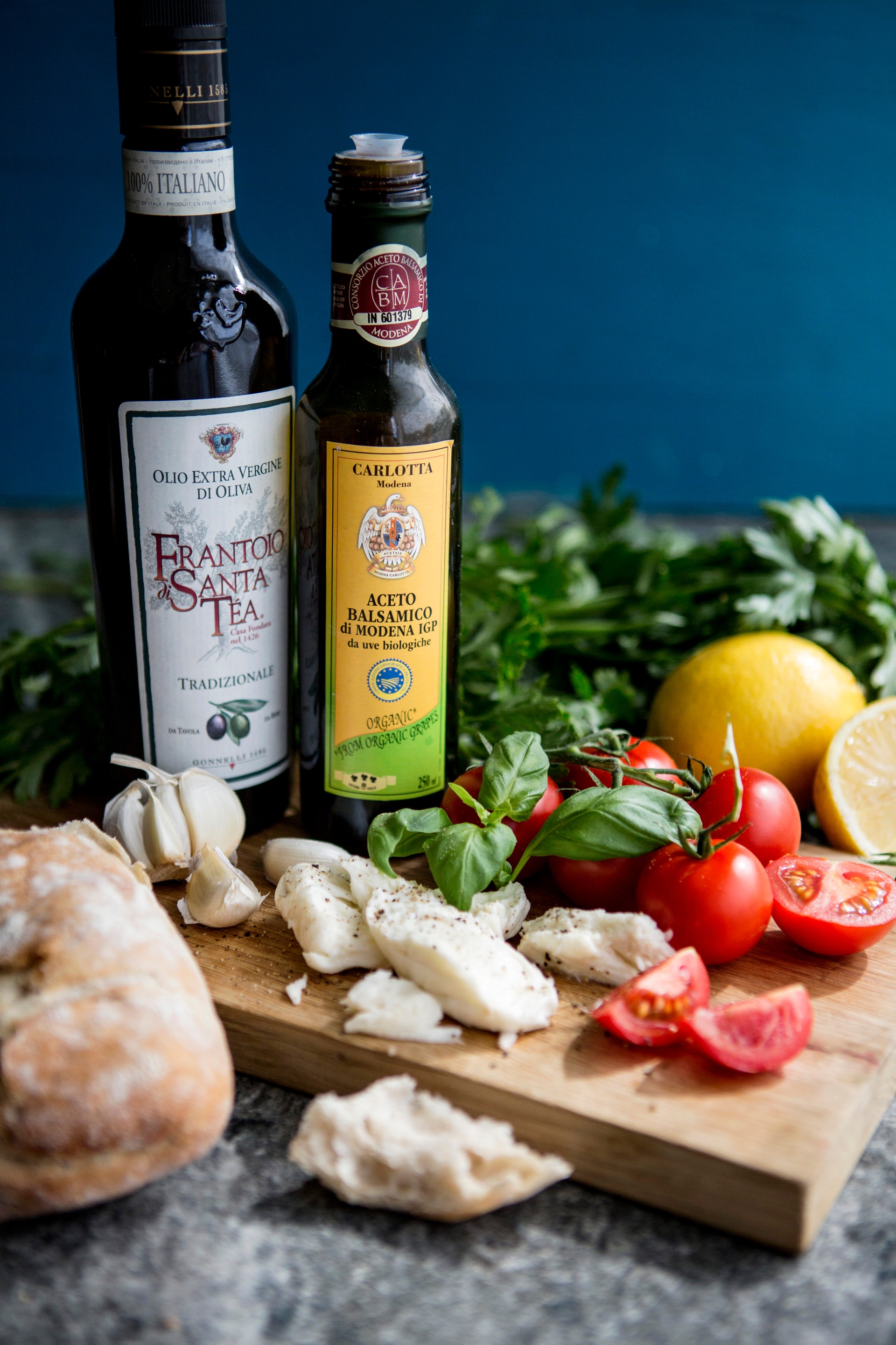 Två flaskor med italiensk olivolja och balsamvinäger på ett skärbräde tillsammans med färska ingredienser: mozzarella, tomater, citron, basilika och vitlök mot en blå bakgrund.