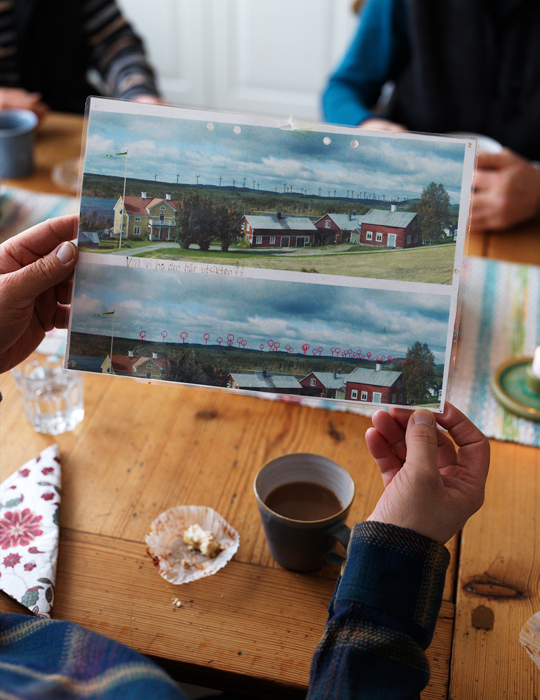 Person håller upp ett fotografi av ett landskap med röda hus och en bro vid ett bord