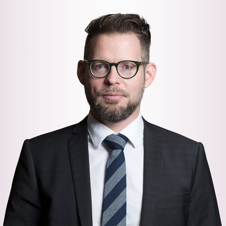 Porträttfoto av en man med glasögon, skägg och mörkt hår, iklädd svart kavaj, vit skjorta och mörkblå randig slips mot ljus bakgrund.