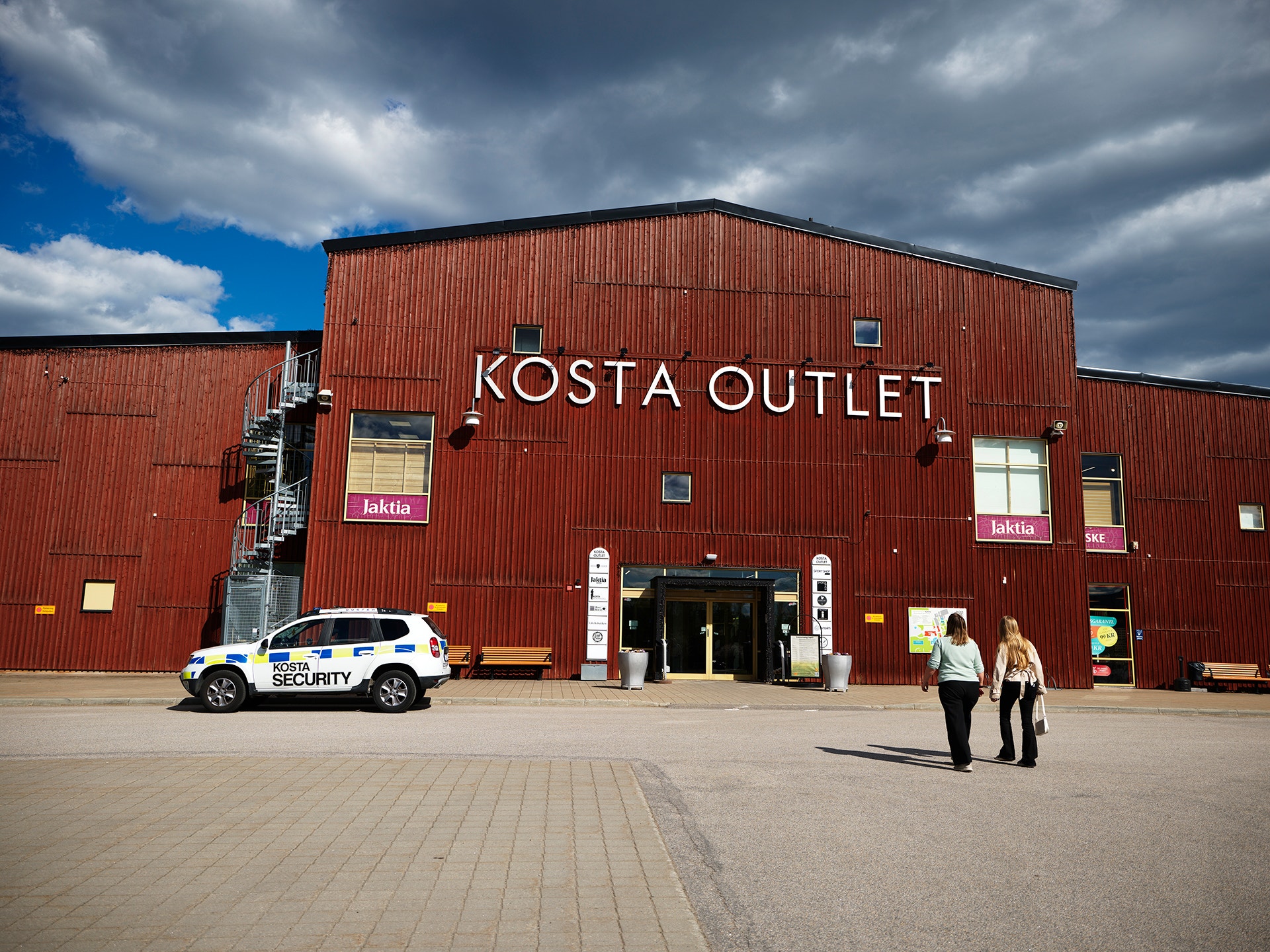 Röd industribyggnad med texten Kosta Outlet på fasaden. En säkerhetsbil och några personer står framför byggnaden på en öppen plats.