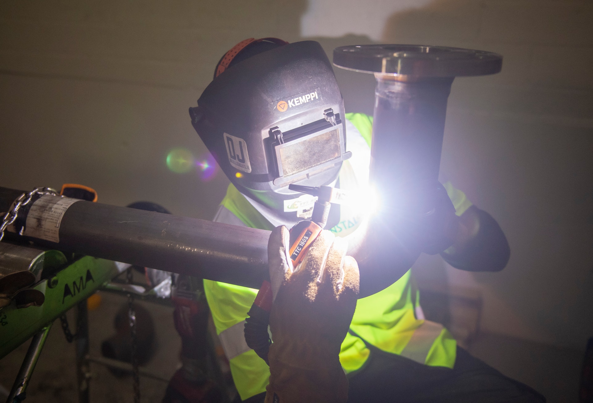 Person i svetshjälm och gul reflekterande väst arbetar med svetsbrännare vid metallarbete.