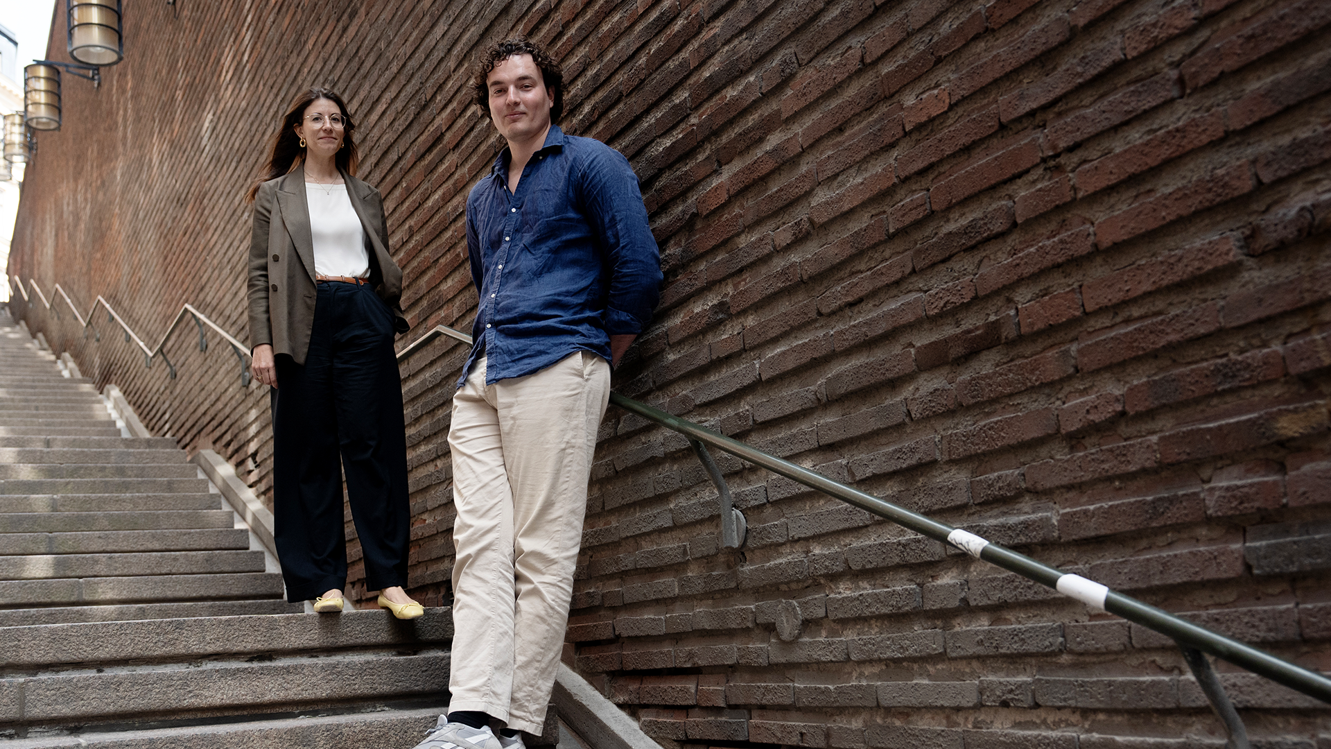 Hanna Andreen och Antonio Melani står på stentrappan vid kungsgatan. De båda tittar in i kameran. Antonio lutar sig med ryggen mot tegelfasaden.