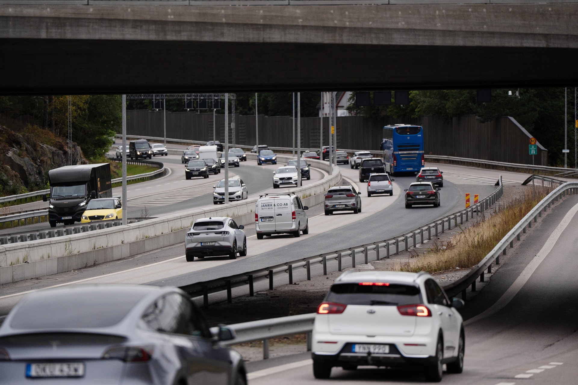 Trafikscen på en motorväg med flera körfält och olika fordon som bilar, lastbilar och bussar under en bro.