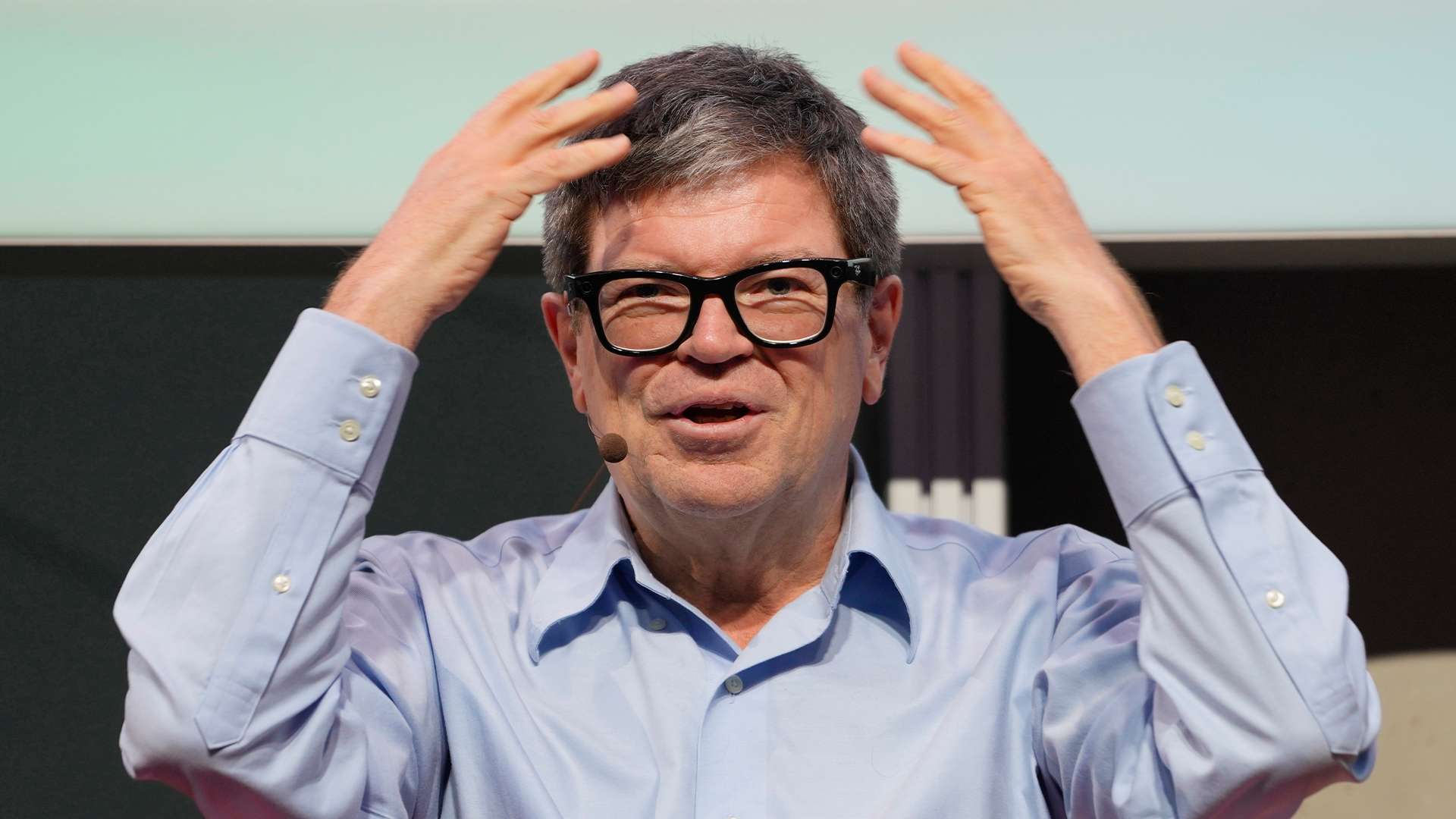 En man med glasögon och ljusblå skjorta med händerna höjda till huvudet i en uttrycksfull gest.