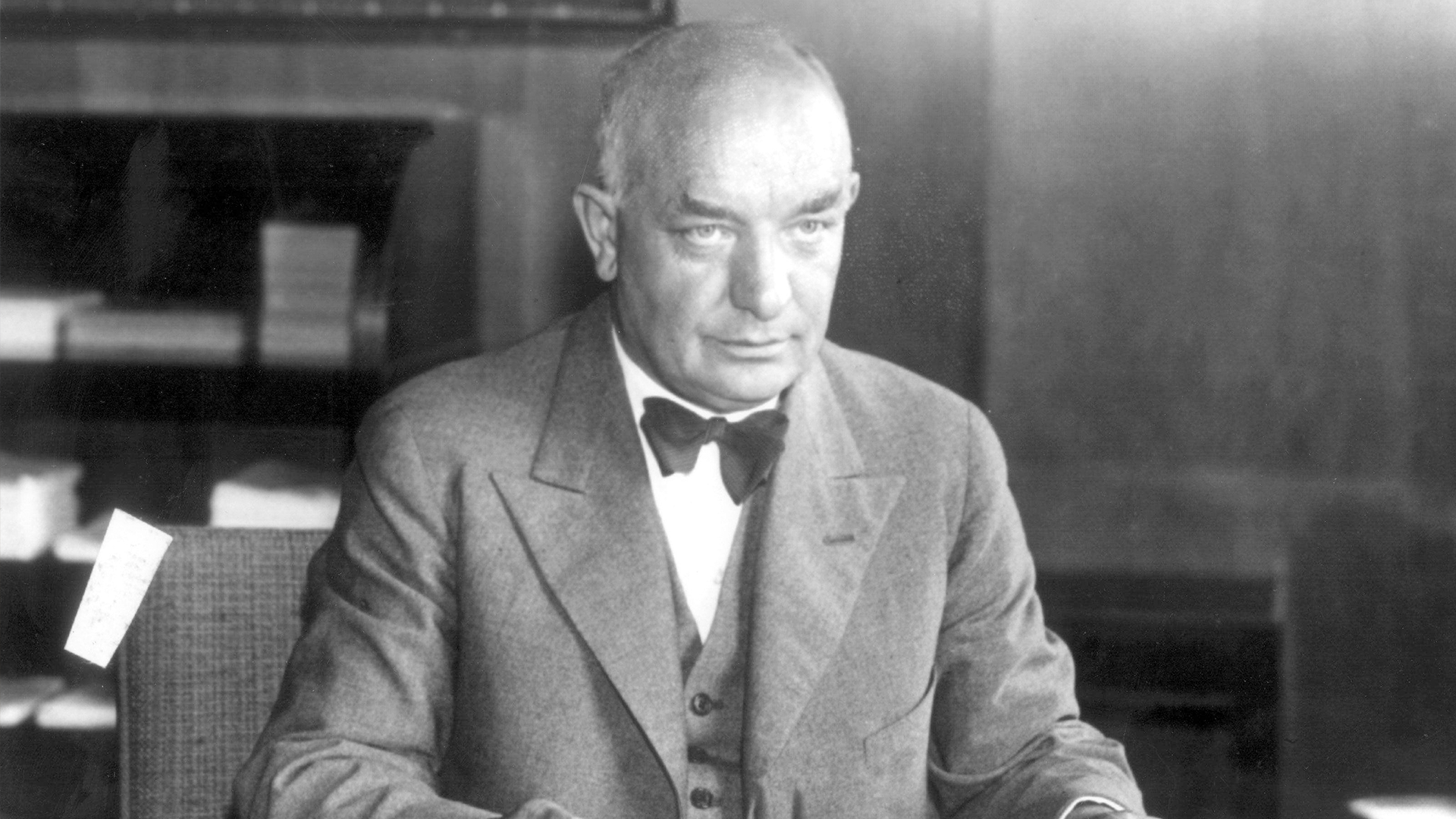 Svartvit porträttfotografi av en äldre man i formell kostym och fluga, sittande vid ett skrivbord.