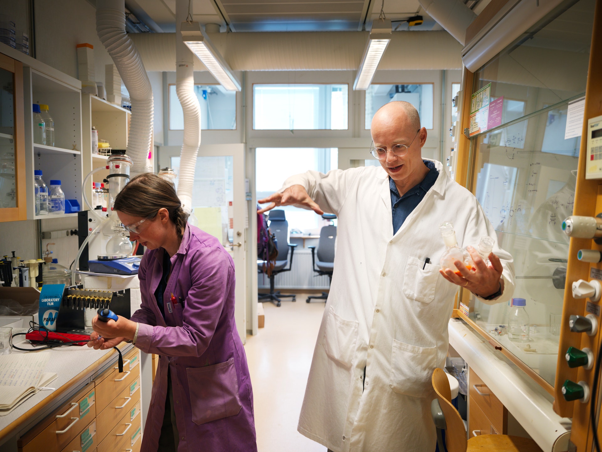 Två forskare arbetar i ett laboratorium. En person i rosa skjorta står vid arbetsbänken till vänster, medan en man i vit laboratoriekittel och blå skjorta står till höger och pekar på något. Laboratoriet är ljust och modernt med fönster, hyllor med utrustning och olika instrument på arbetsbänkarna.