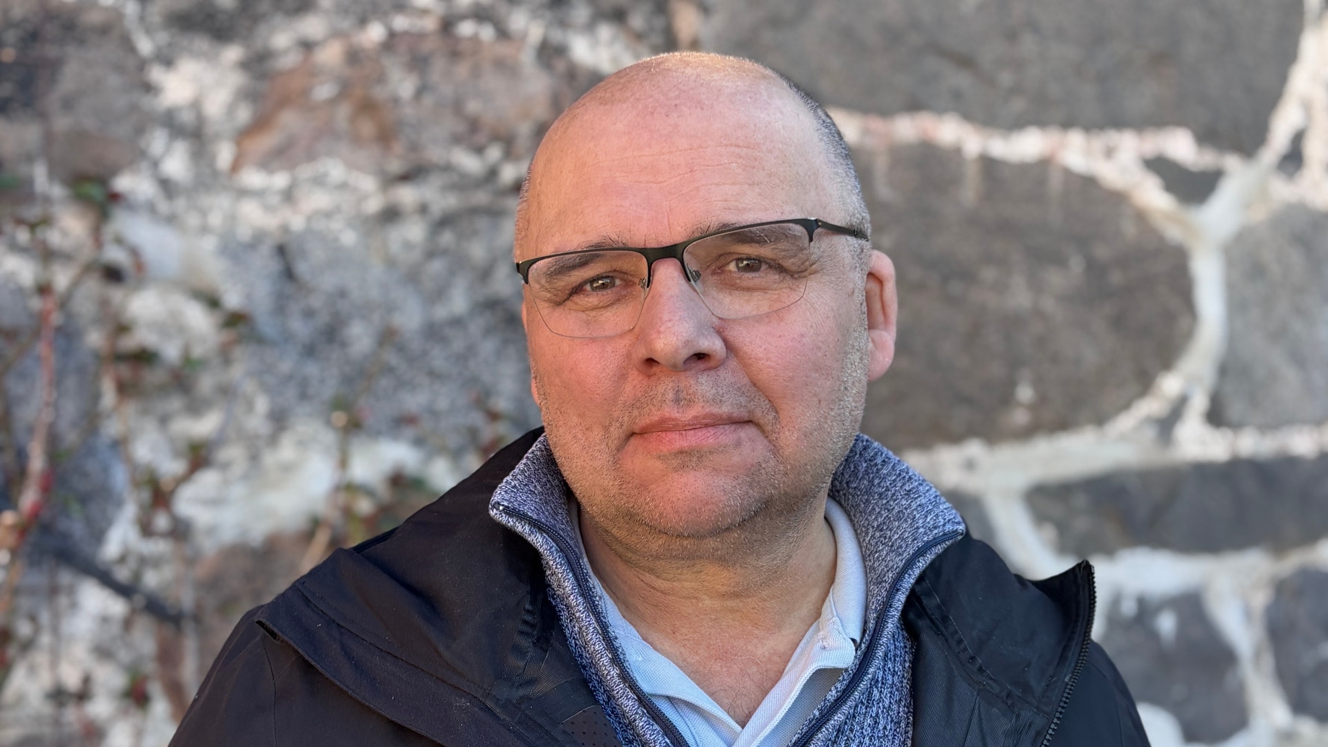 Porträtt av en man med glasögon och mörk jacka framför en tegelvägg.