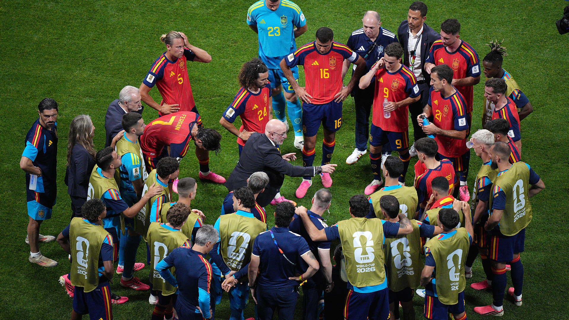 Fotbollslag i röda och mörkblå uniformer samlade på gräsmattan med tränare och stödjepersonal för ett möte under matchen.