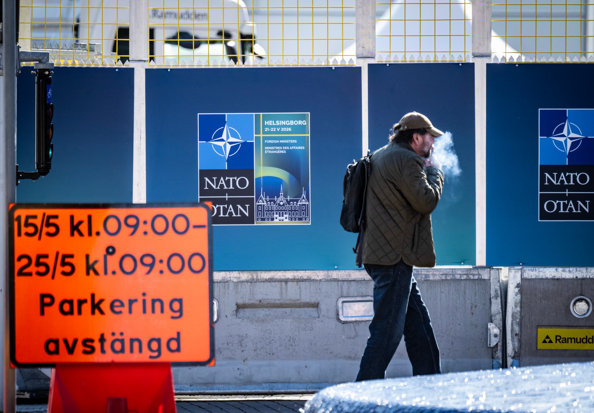 En man går förbi en blå vägg med NATO-logotyper inför ett NATO-möte i Helsingborg. Snö ligger på marken och en orange parkeringsskylt syns i förgrunden.