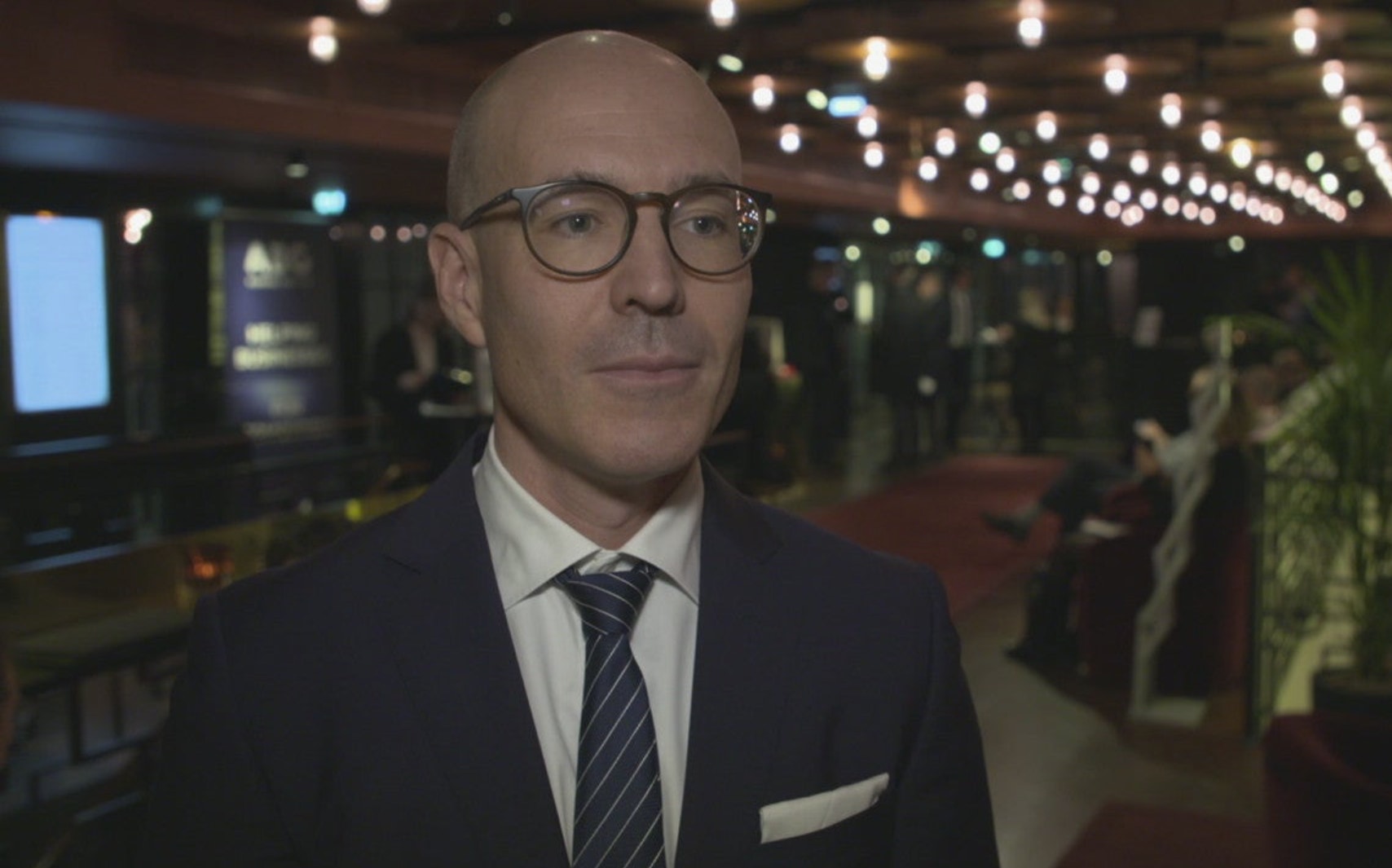 En man i kostym och glasögon står i en inomhusmiljö med dekorativ belysning i bakgrunden.