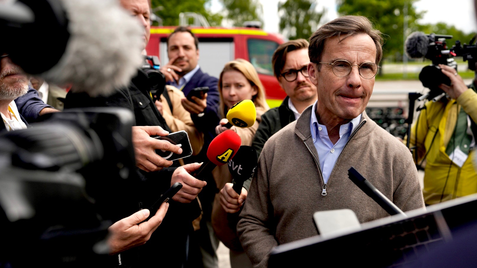 En man i brun tröja talar vid en presskonferens omgiven av journalister med mikrofoner och kameror. En röd brandbil syns i bakgrunden.