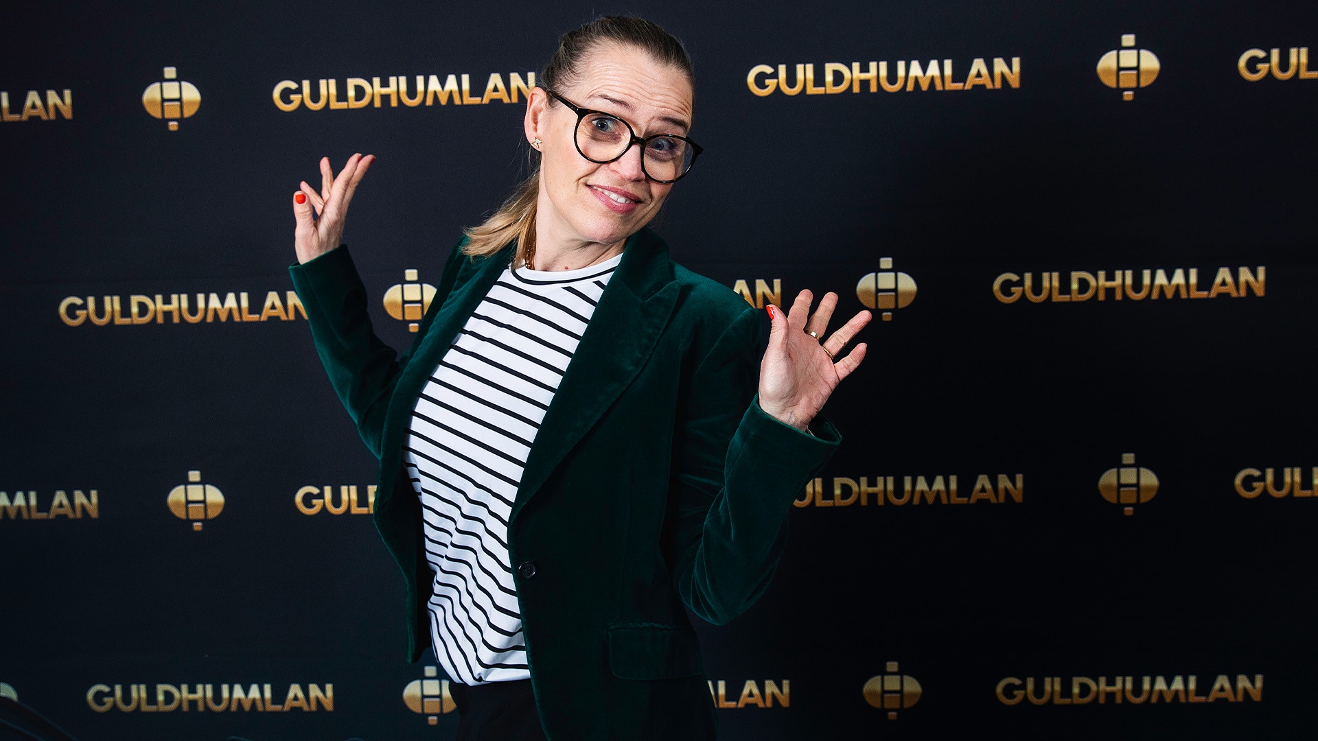 Person i grön kavaj och randig tröja poserar glatt framför en Guldhumlan-bakgrund.