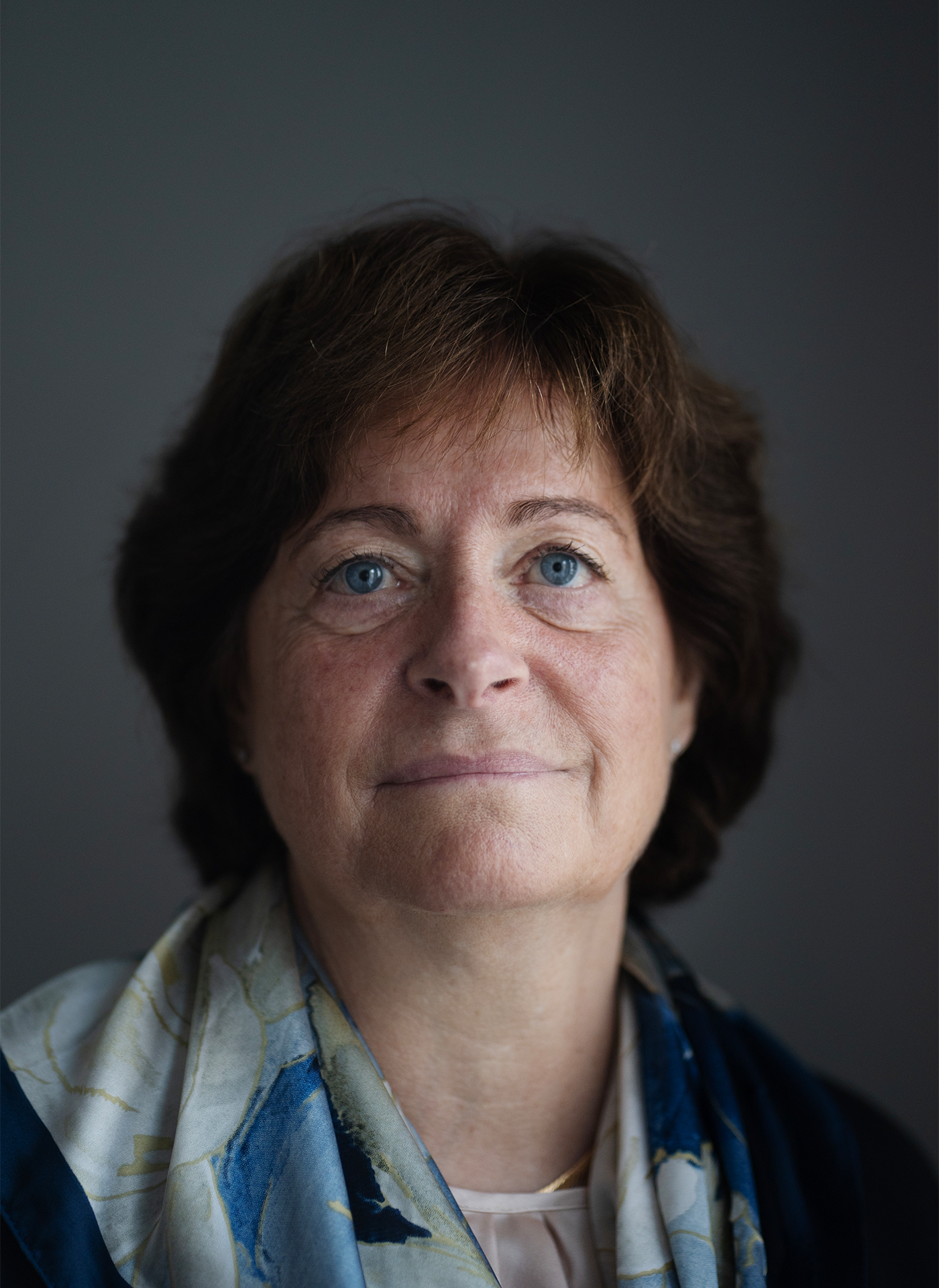 Gunilla Osswald tittar in i kameran. Hon står i vardagliga kläder mot en grå bakgrund.