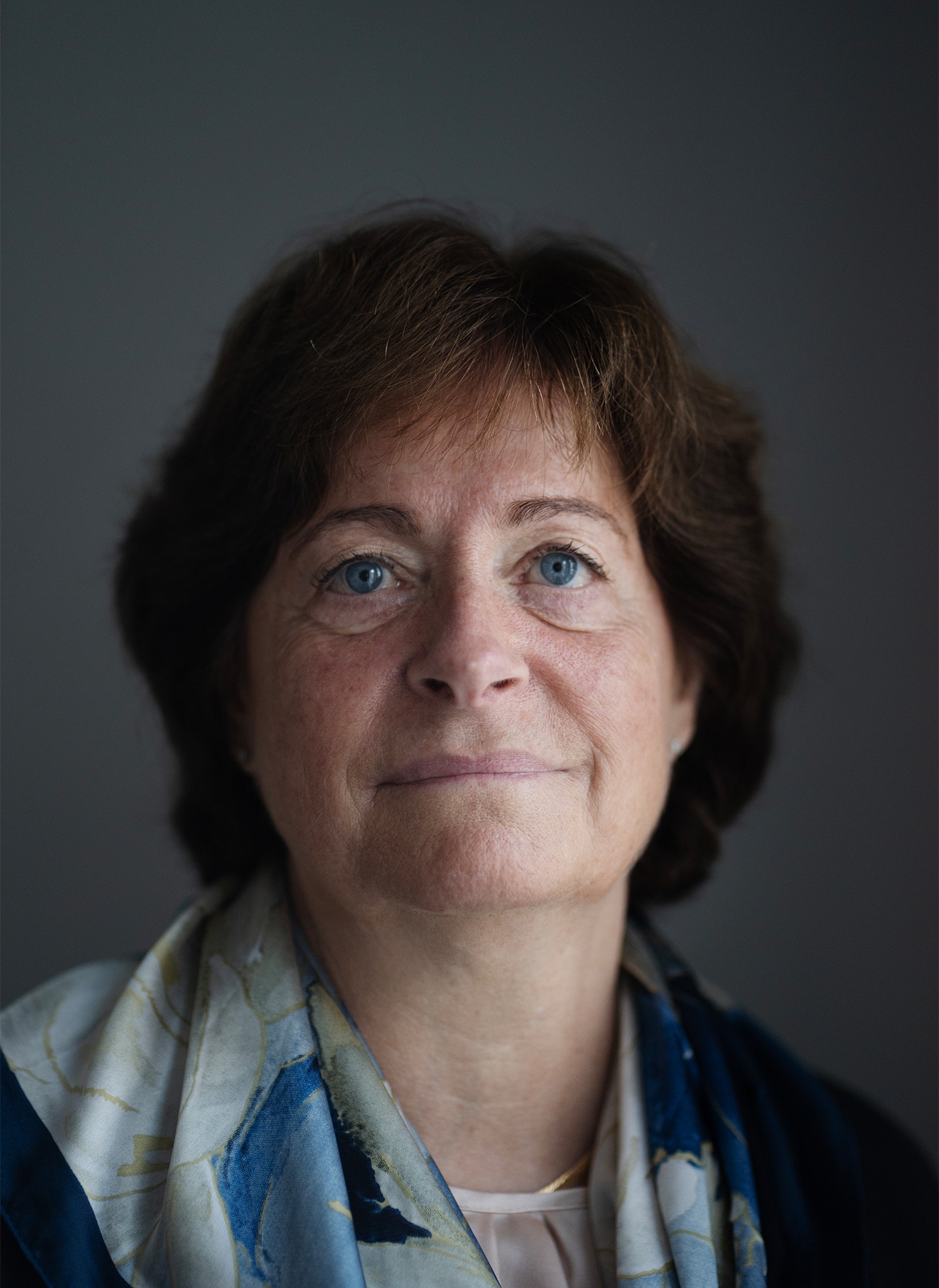 Gunilla Osswald tittar in i kameran. Hon står i vardagliga kläder mot en grå bakgrund.