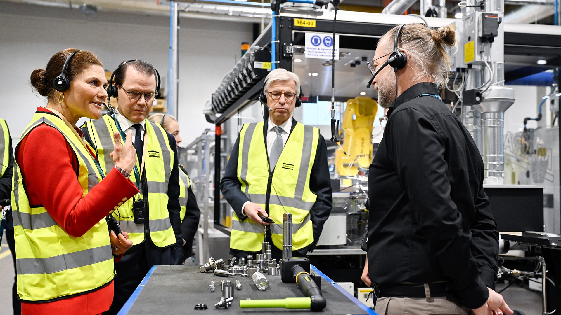 Fyra personer i gula säkerhetsvästor står omkring en industriell maskin i en fabrik. En person bär hörlurar och presenterar maskinen för de andra.