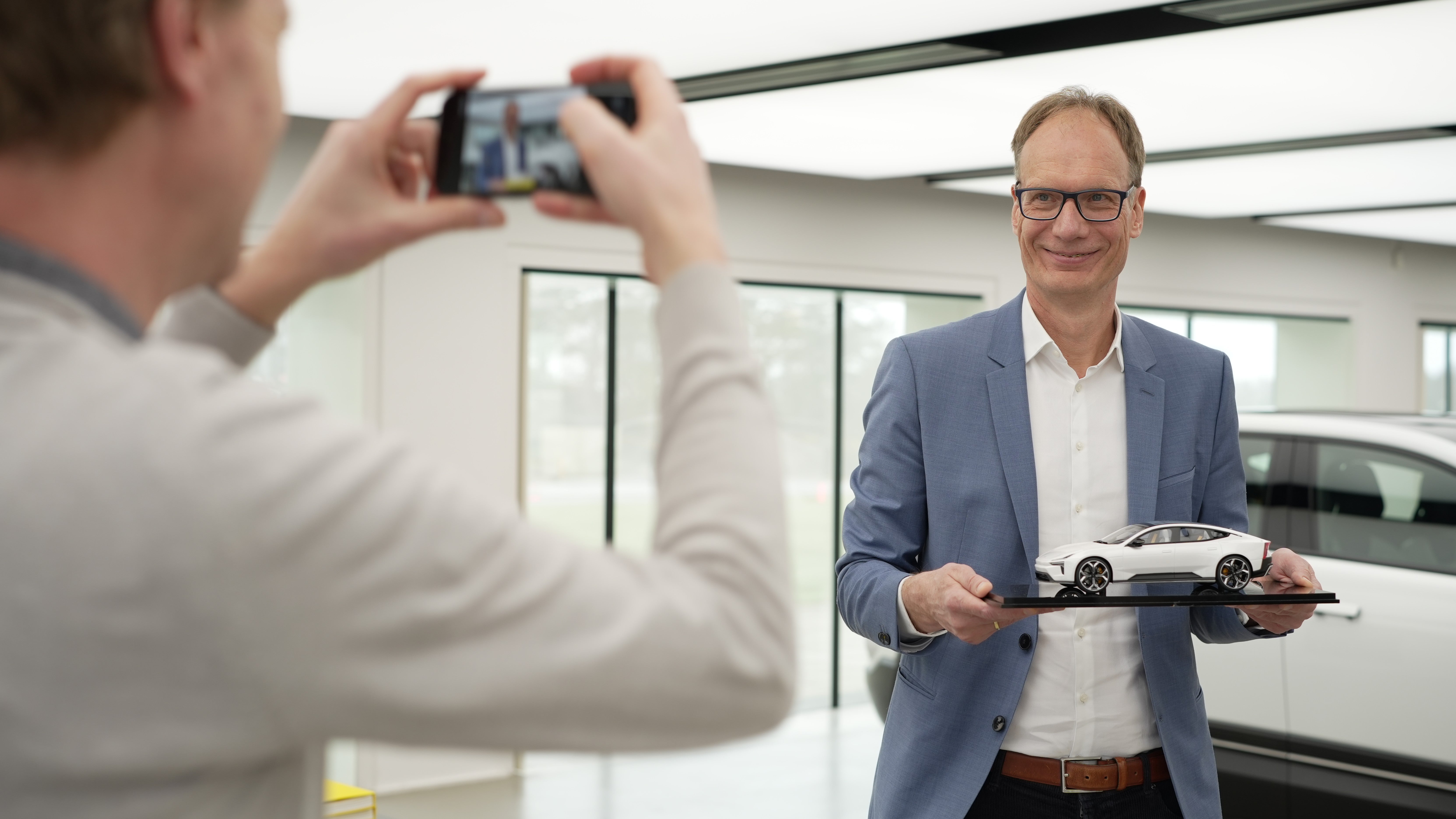 Man i blå kavaj håller en vit bilmodell medan en annan person fotograferar honom med en smartphone.