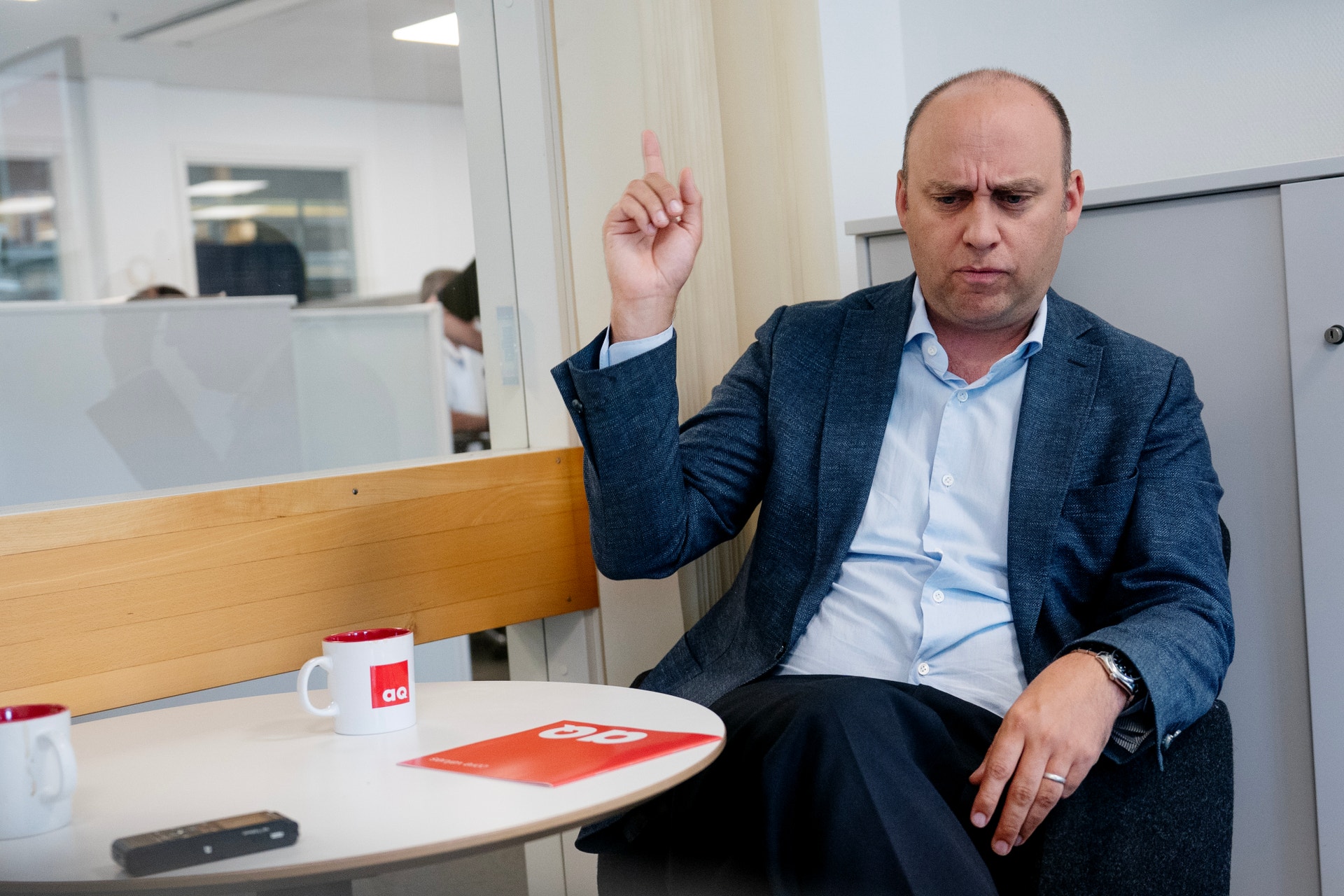 Man i mörkblå kavaj sitter vid ett skrivbord på ett kontor och höjer sitt högra finger medan han talar.
