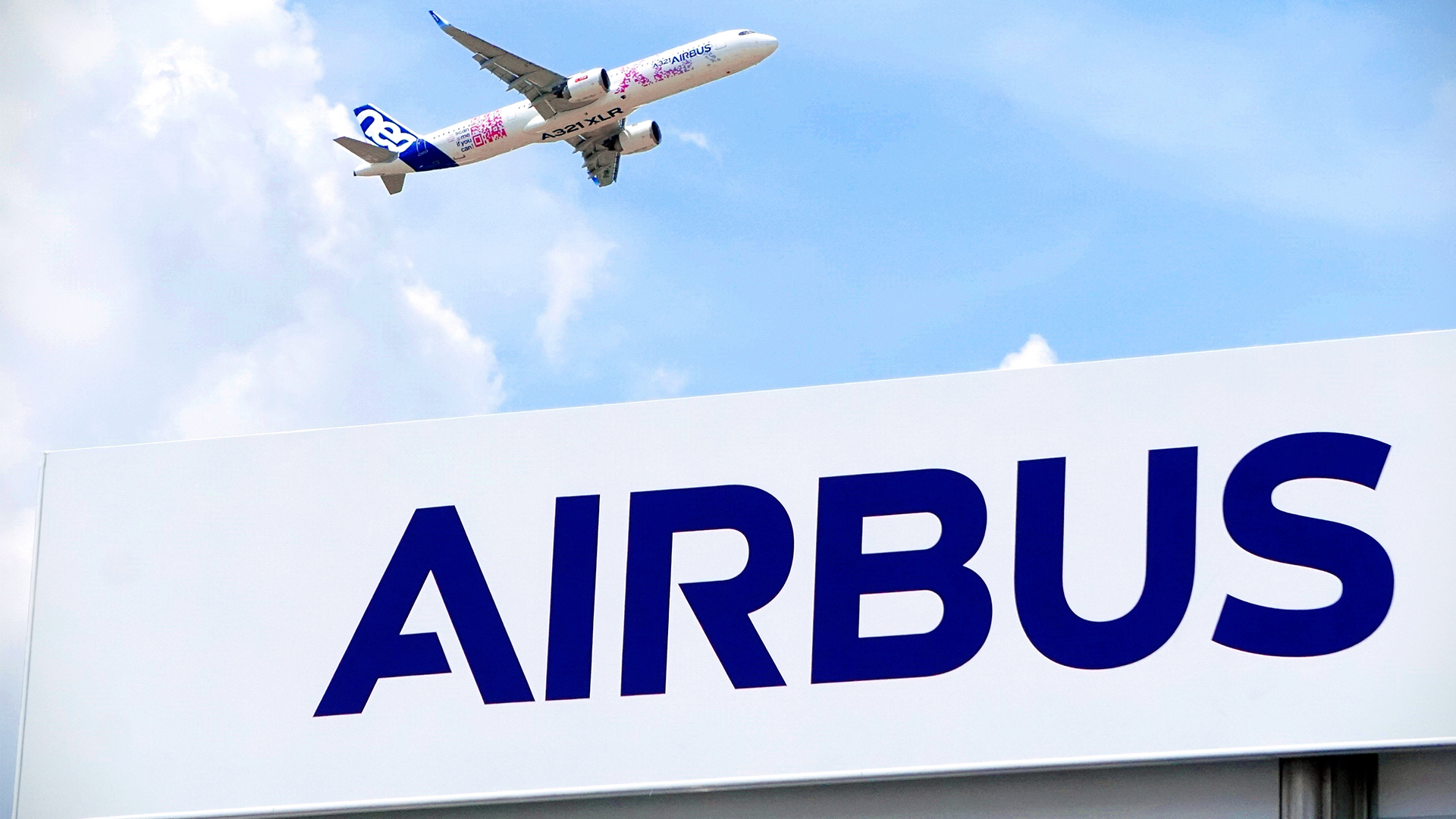 Ett Airbus-flygplan flyger över en stor skylt med texten Airbus mot en blå himmel med moln.