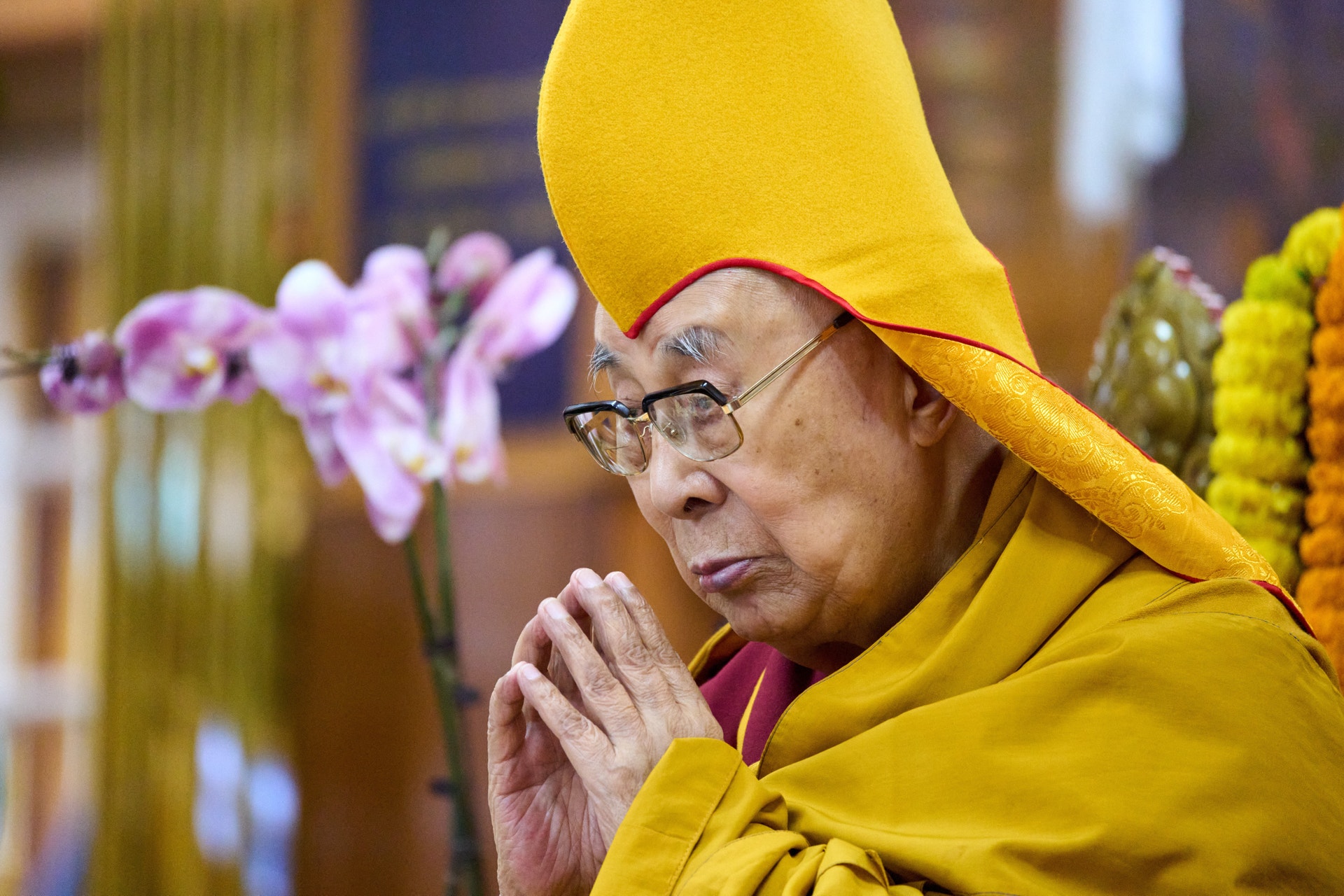 En person i gul munkdräkt och gul mössa sitter i en meditativ hållning med händerna sammanslagna framför sig. Färgglada blommor syns i bakgrunden.