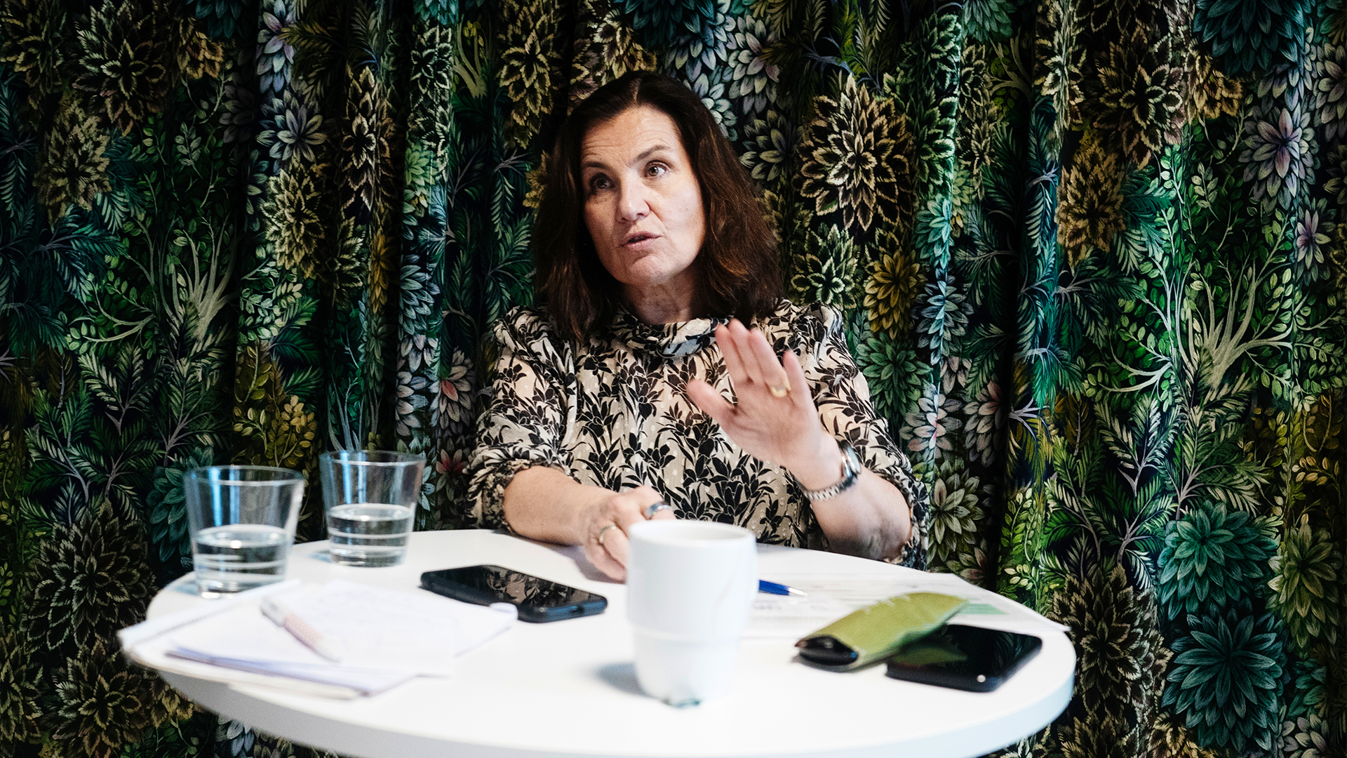 Ann Carlsson Meyer står och pratar vid ett barbord. På bordet framför henne står det kaffe, vatten, anteckningsblock, glasögon och två mobiltelefoner.