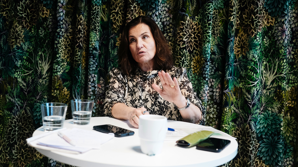 Ann Carlsson Meyer står och pratar vid ett barbord. På bordet framför henne står det kaffe, vatten, anteckningsblock, glasögon och två mobiltelefoner.