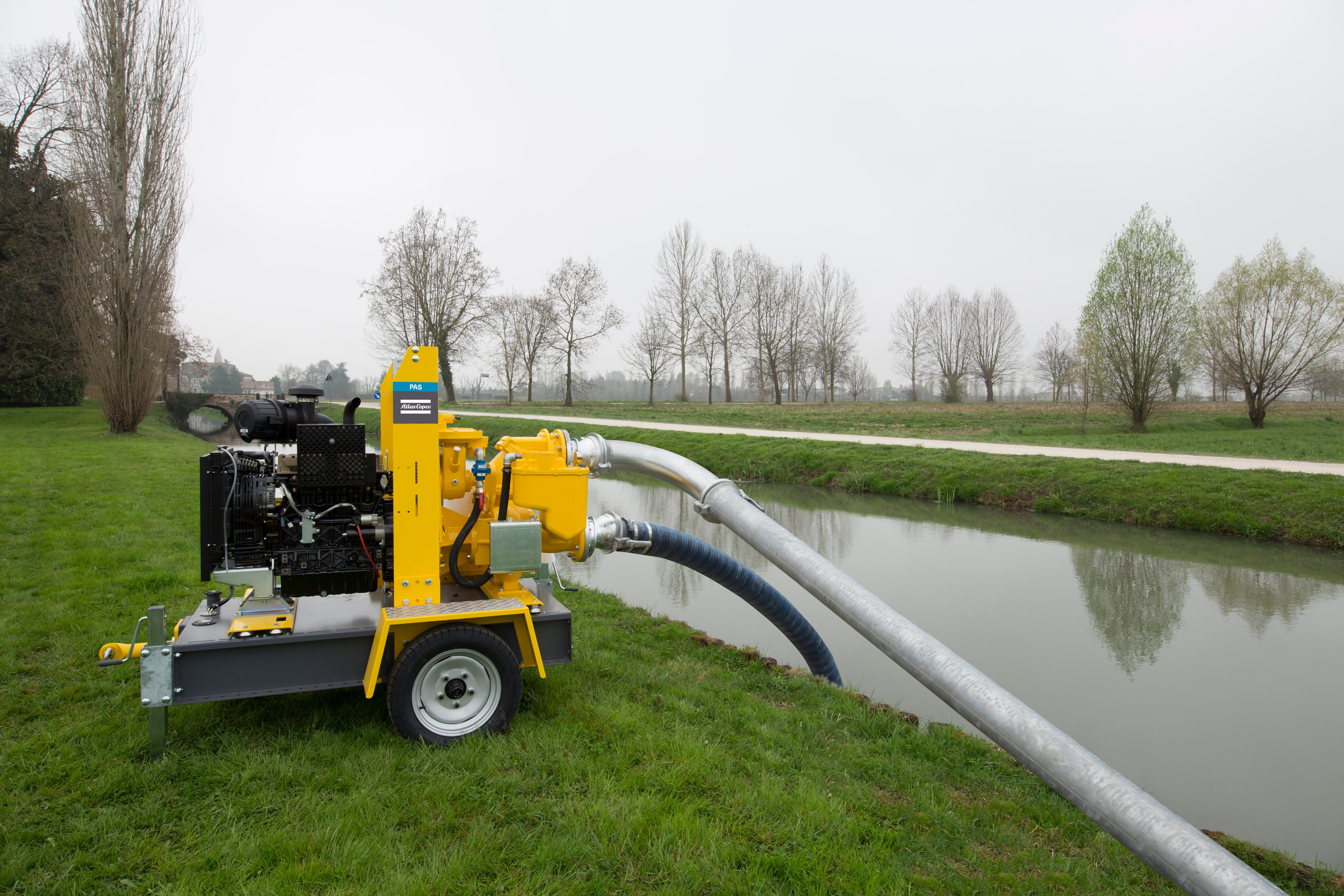 Gul pumpstation på hjul placerad på en äng bredvid en kanal, med ett rör som sträcker sig ner i vattnet för avvattning.