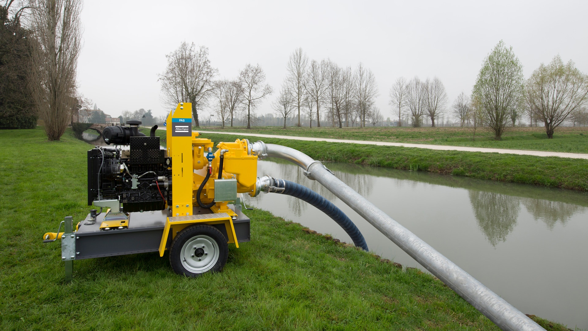 Gul pumpstation på hjul placerad på en äng bredvid en kanal, med ett rör som sträcker sig ner i vattnet för avvattning.