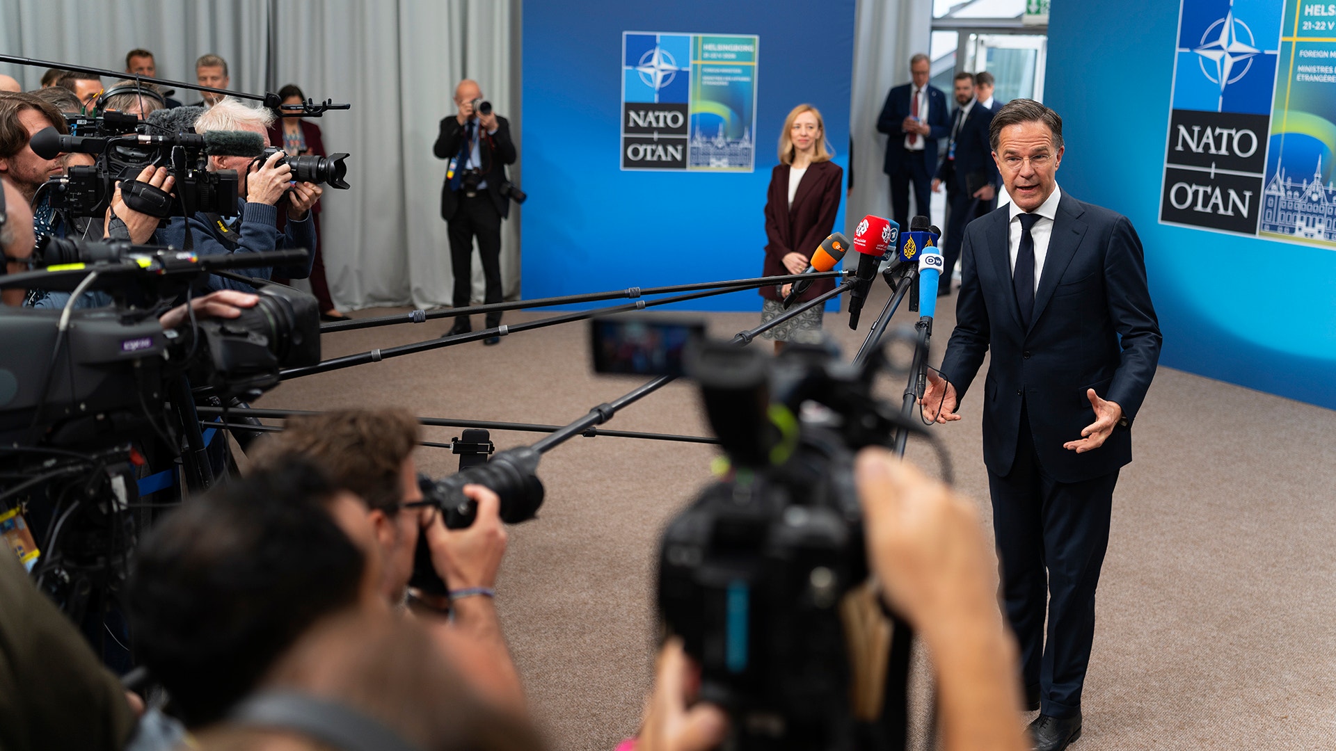 En man i mörk kostym går mot en grupp journalister och TV-kameror under en presskonferens. NATO-loggor syns på blå bakgrund bakom honom.