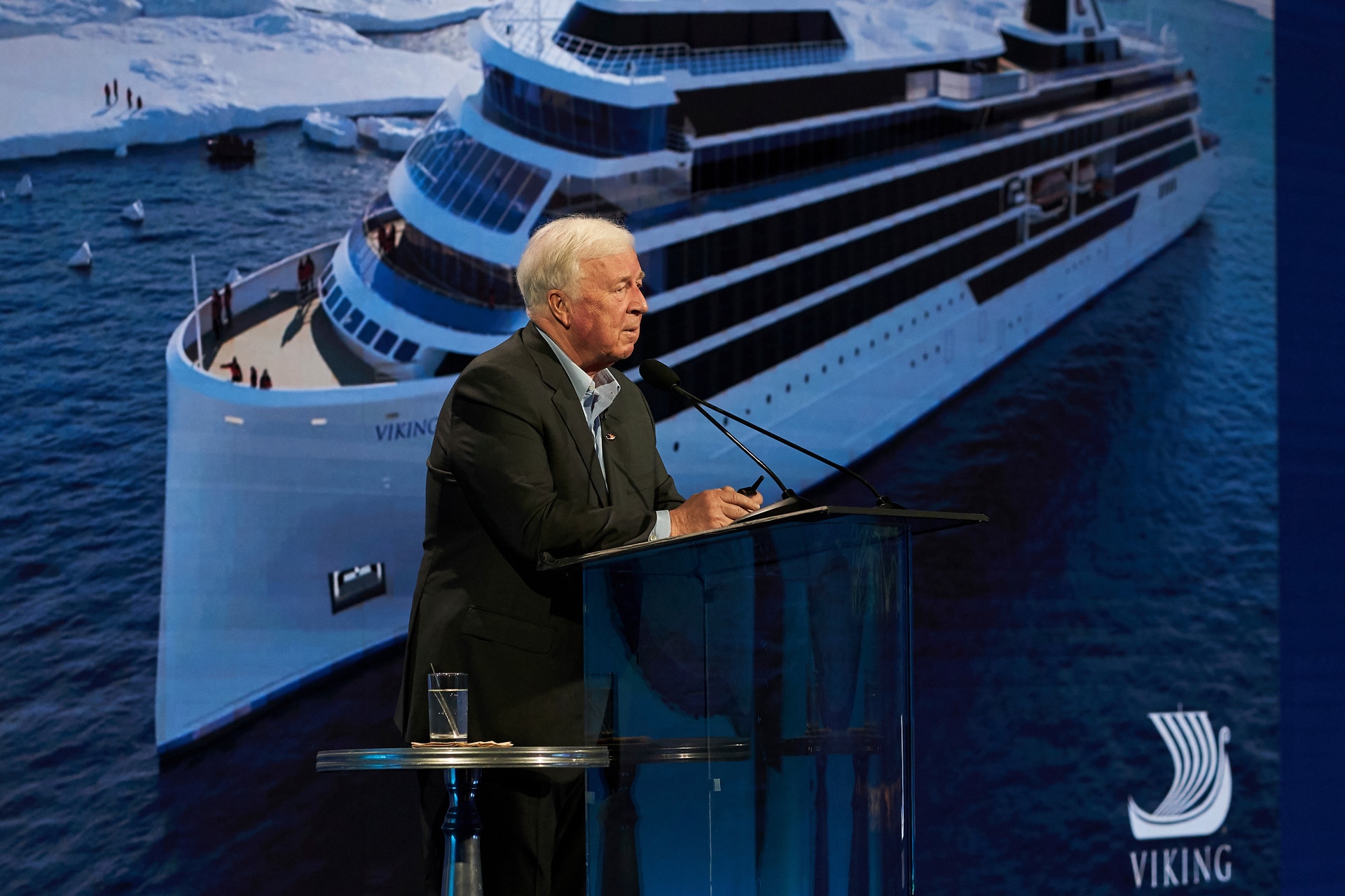 En äldre man i mörk kostym talar vid ett podium. I bakgrunden syns ett stort modernt kryssningsfartyg på havet.