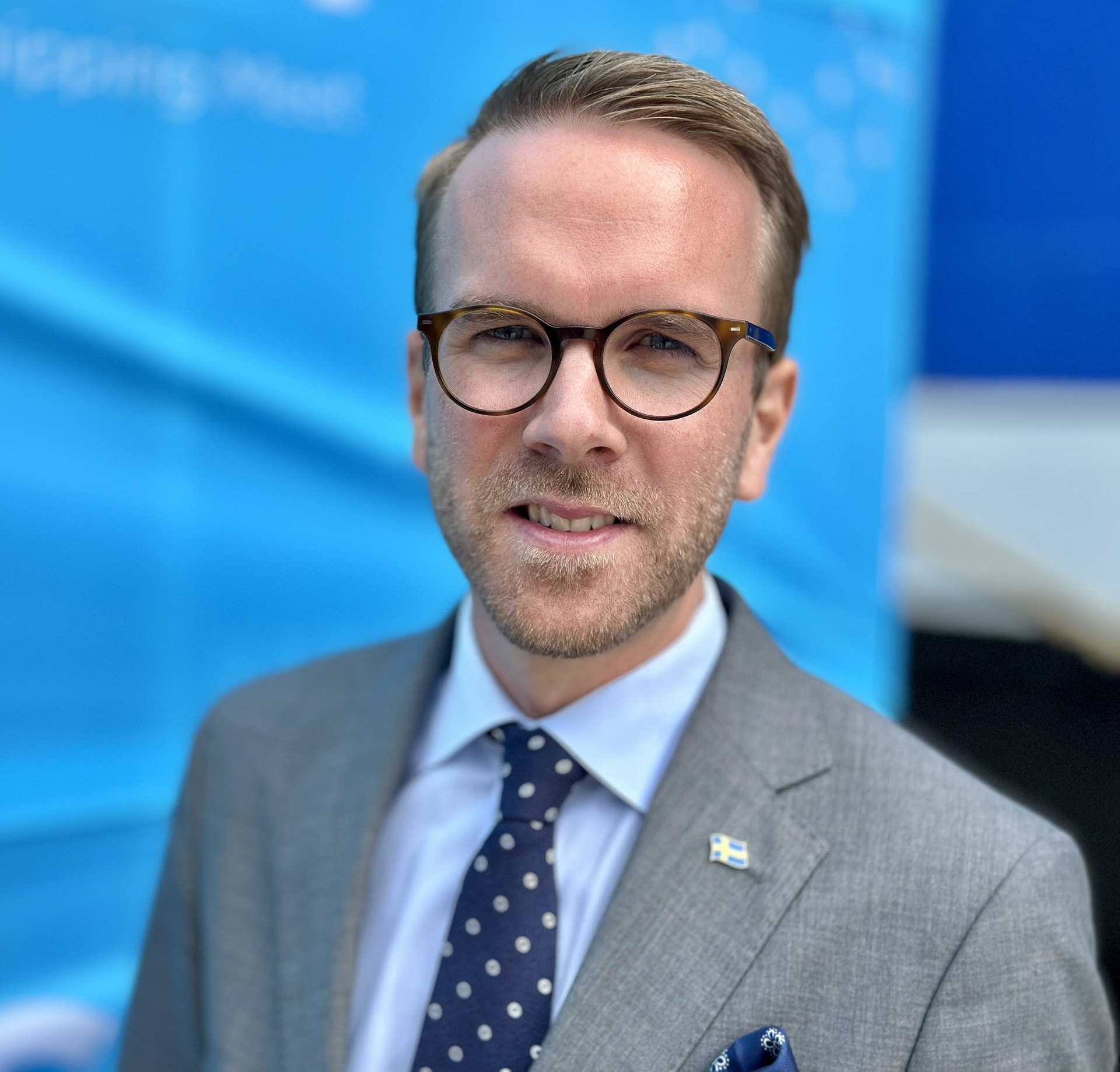 Porträtt av en man med glasögon, grå kostym och blå slips mot blå bakgrund.