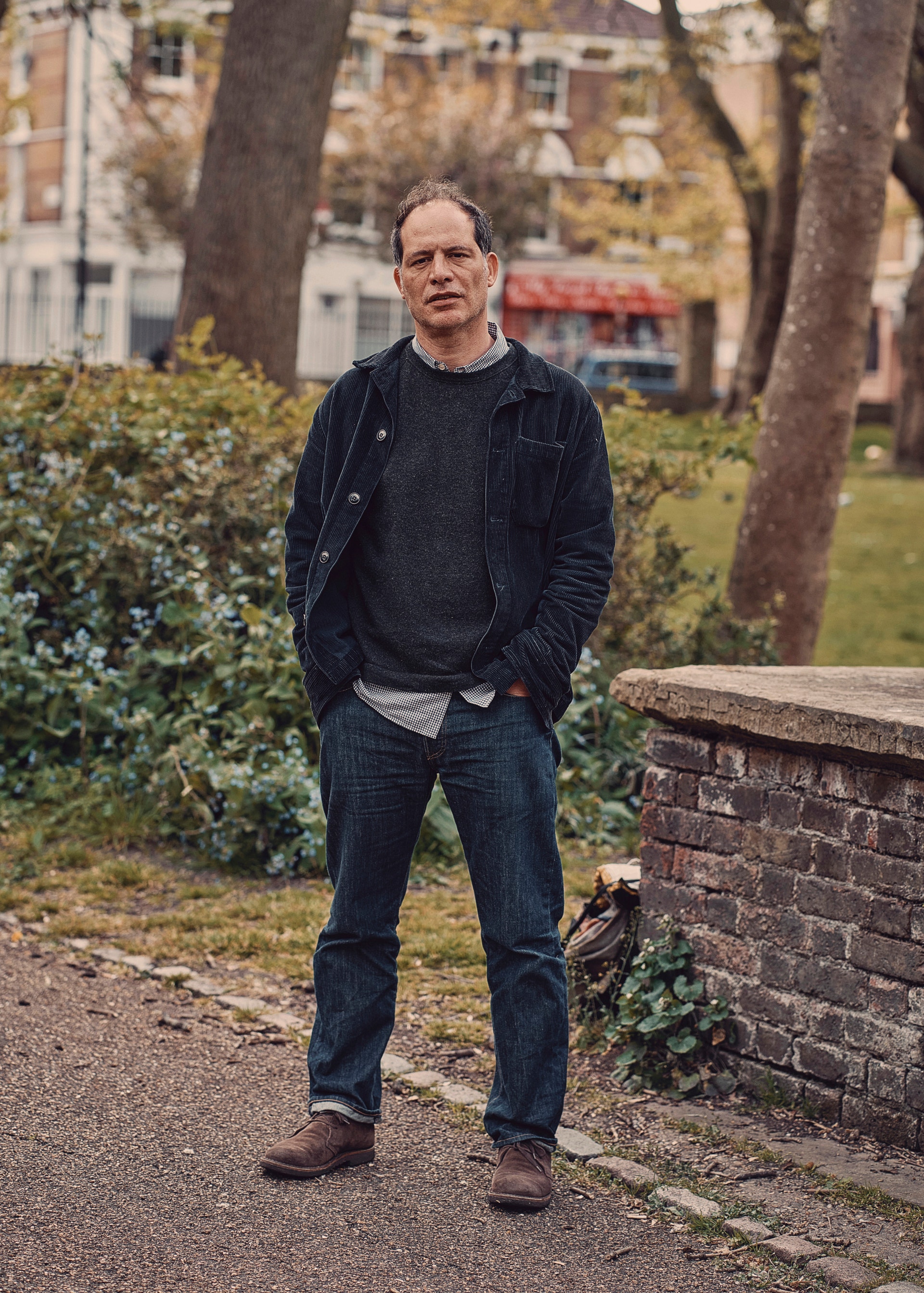 Porträtt av en man med glasögon som står i en park omgiven av träd och gröna buskar. Han bär en mörk cardigan och jeans.