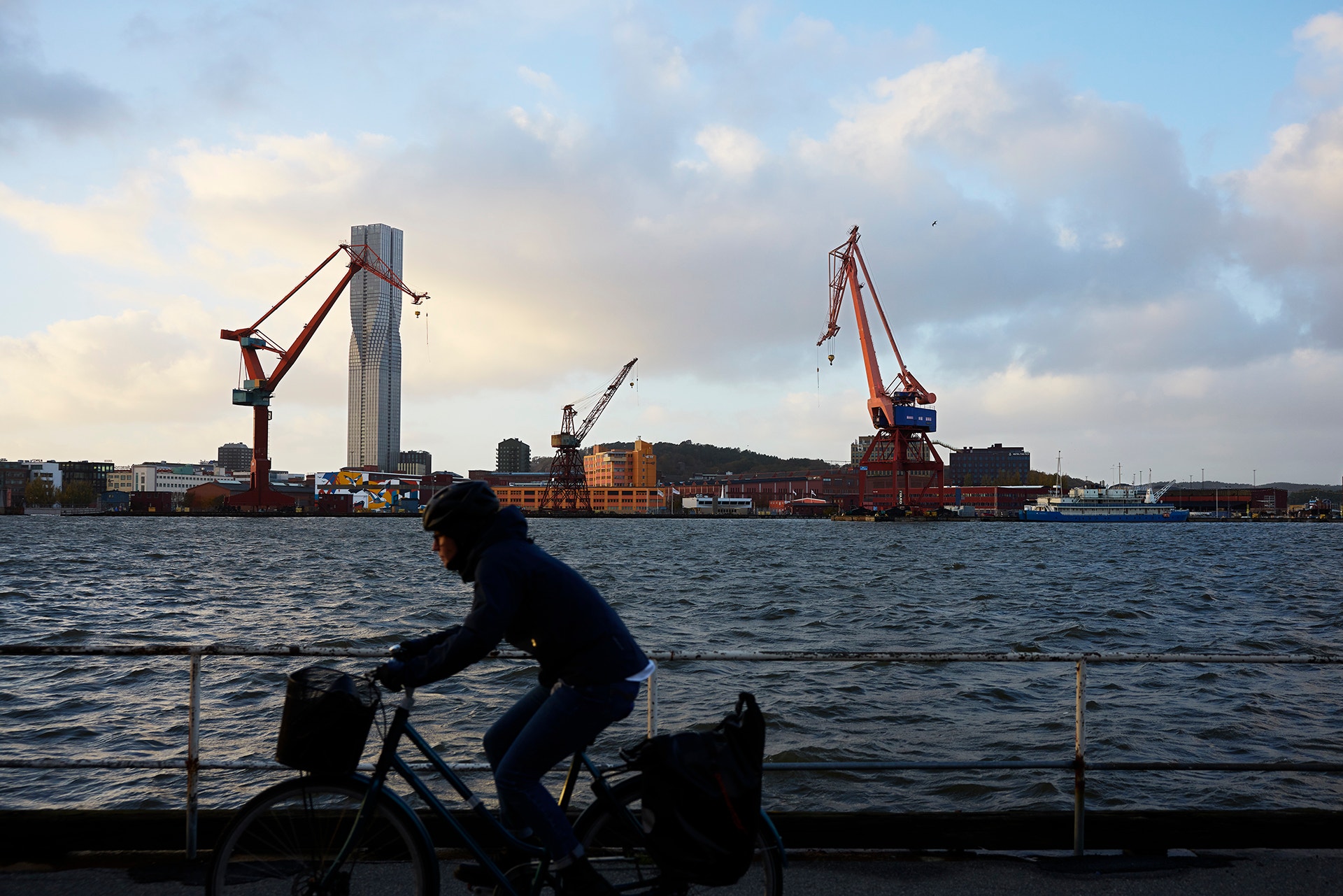 En cyklist i siluett cyklar längs vattnet i Göteborg, med hamnkranar och en hög skyskrapa i bakgrunden.