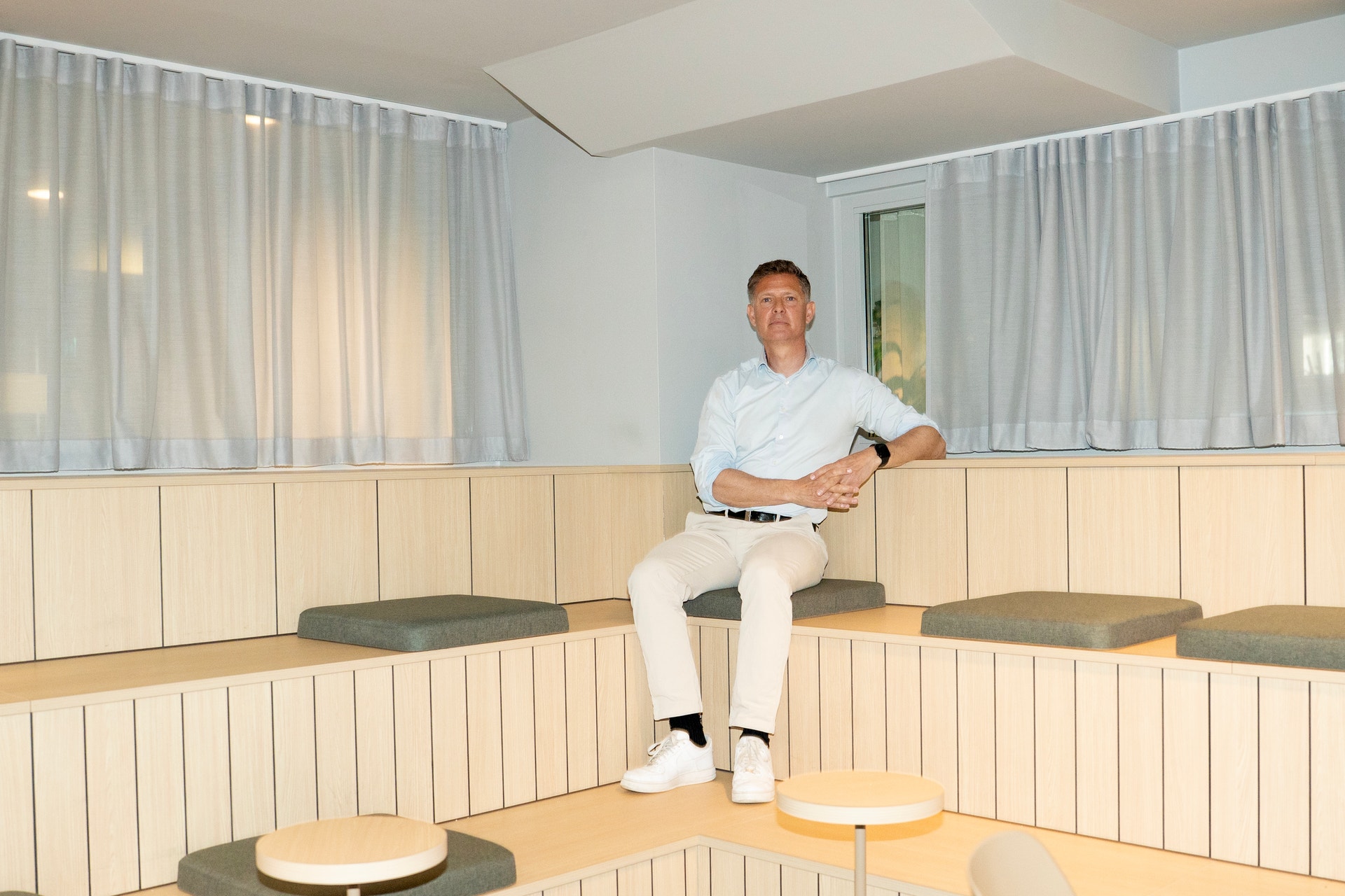 Man i vit skjorta sitter på en träbänk i ett modernt rum med ljusa väggpaneler och stora fönster.