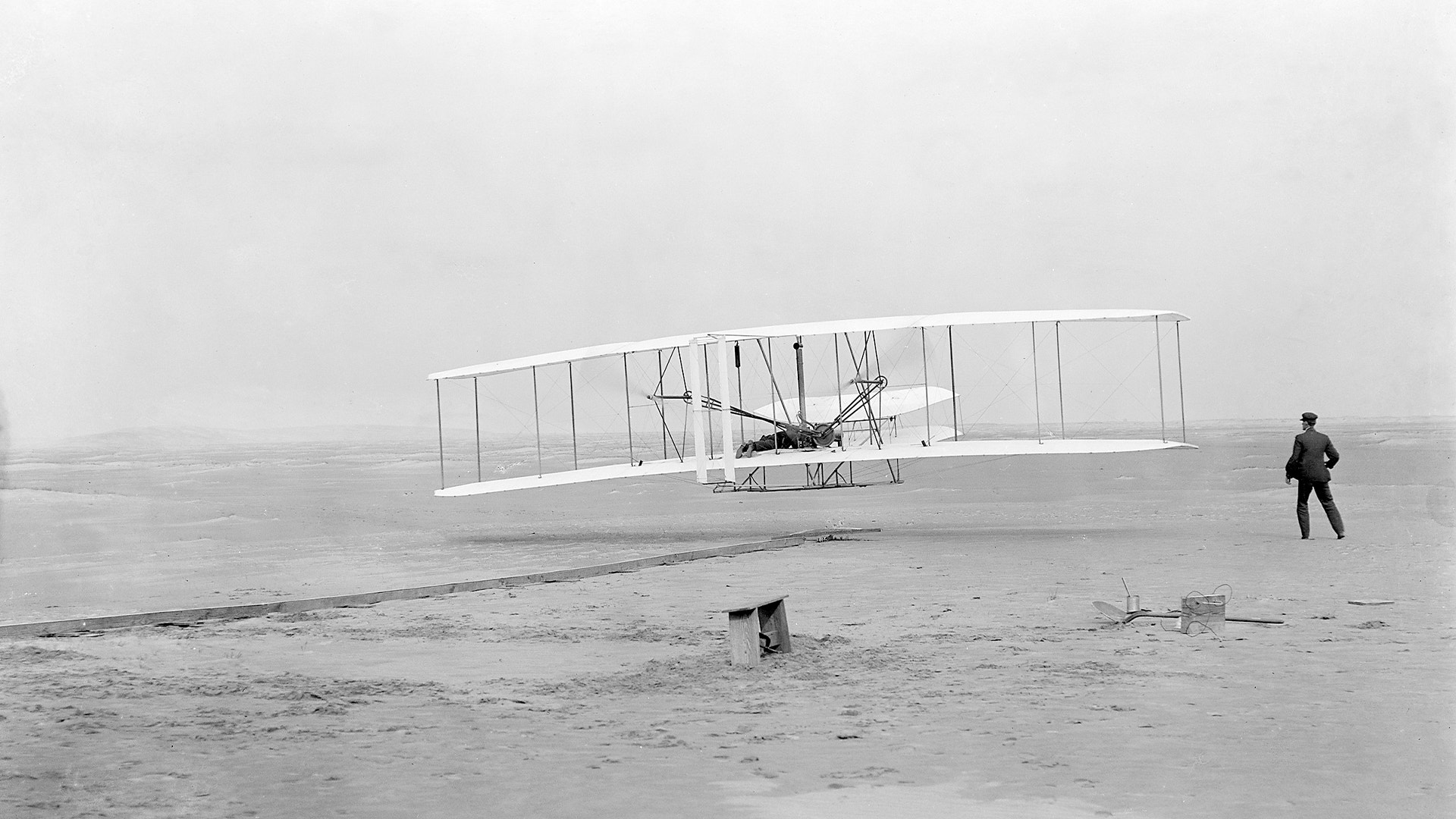 Historiskt flygplan med stora vingar på en sandstrand. En person står i bakgrunden. Svartvit fotografi från flygplanets tidiga utveckling.