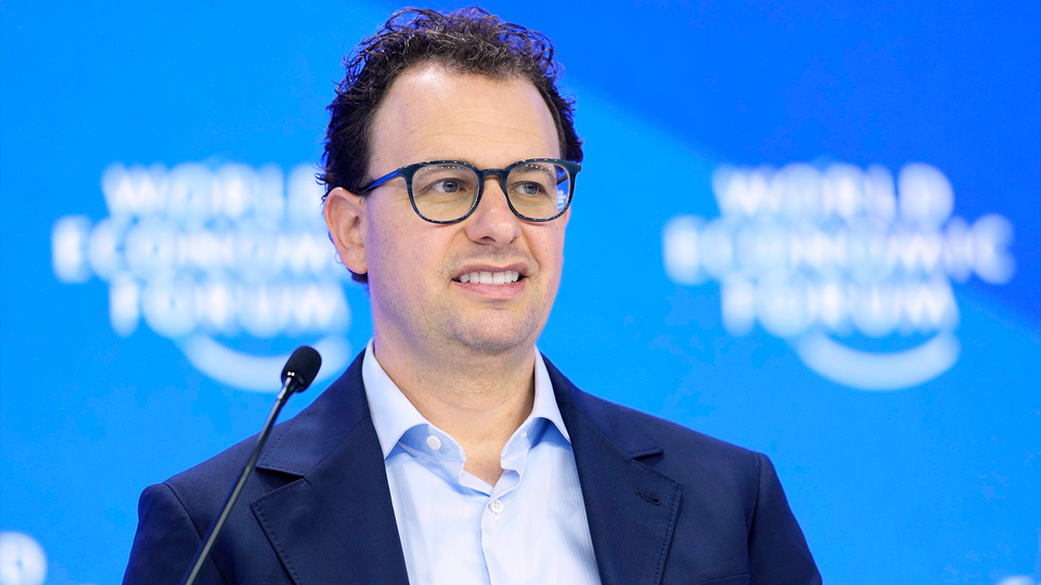 Man med glasögon som talar vid en mikrofon framför en blå bakgrund på en konferens.