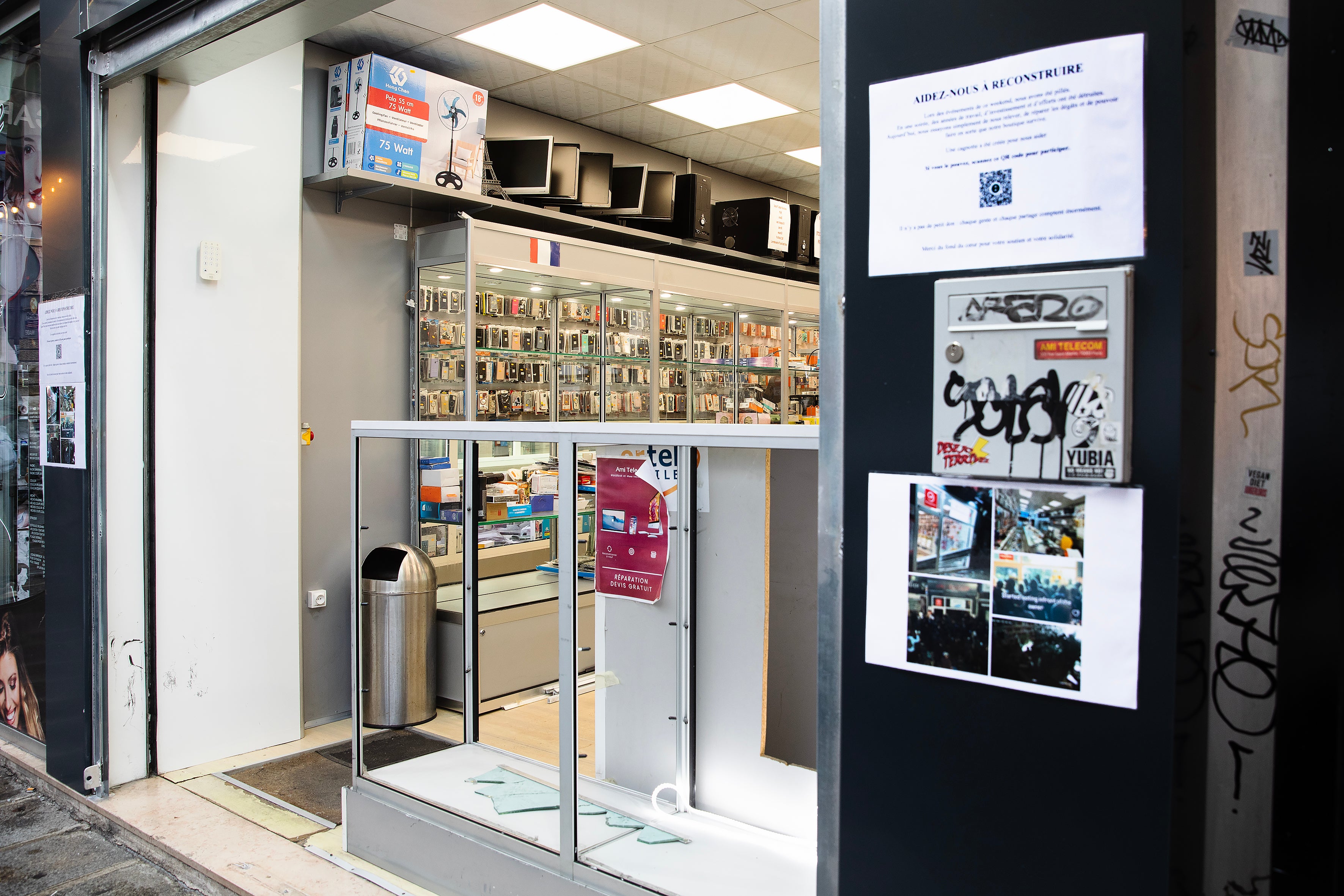 Entrén till en teknikbutik med glasmonter och elektronik. Bredvid dörren sitter affischer och foton på en mörk vägg med klotter.