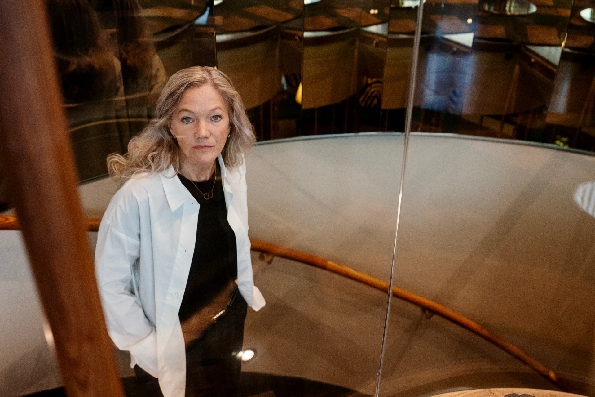 Porträtt av en kvinna med ljust hår, klädd i vit skjorta och svart tröja, som står framför stora glasfönster i en modern byggnad.