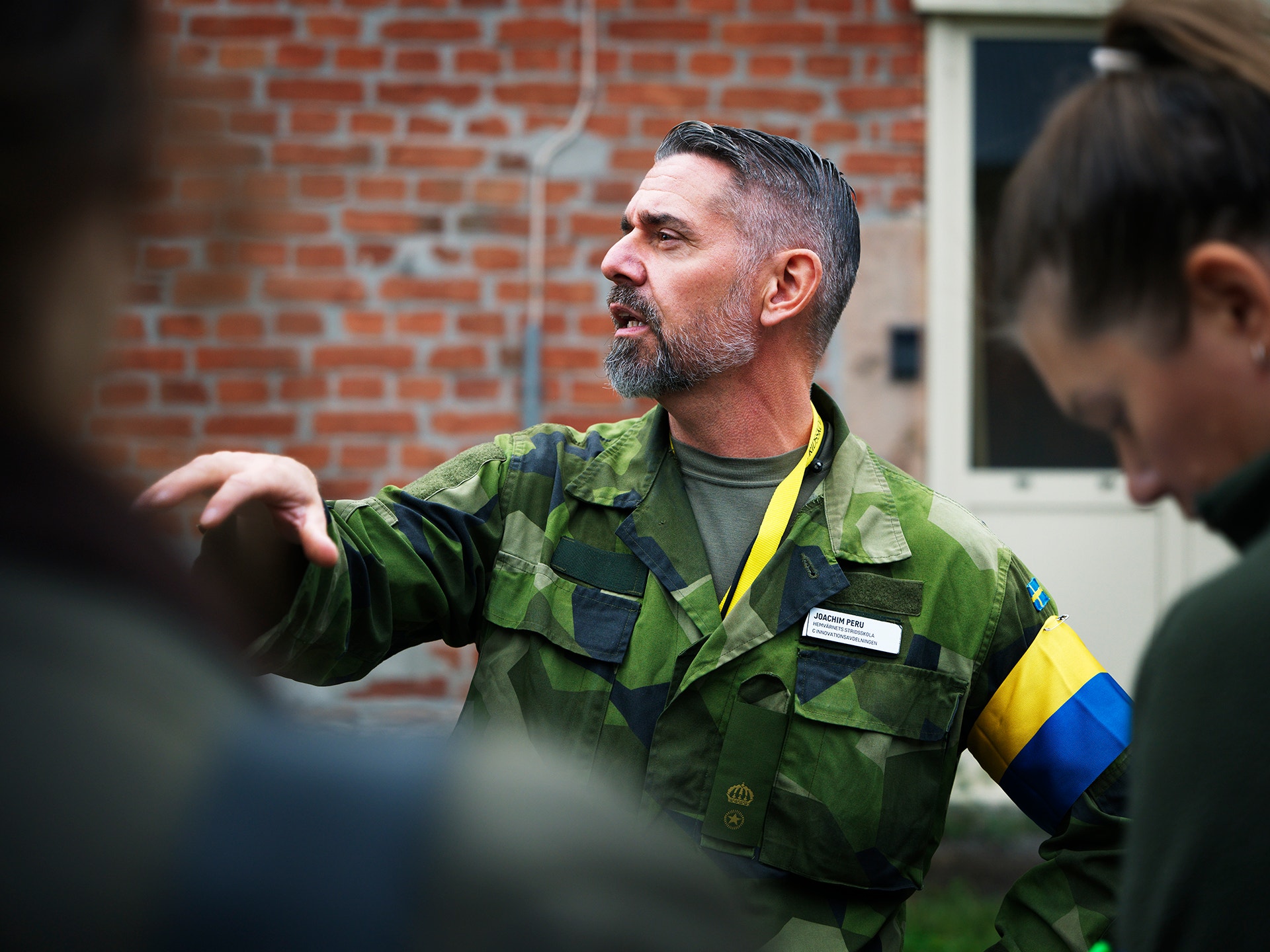 En man i militär kamouflageuniform med svensk flagga på armen pratar och gestikulerar utomhus, omgiven av andra personer.