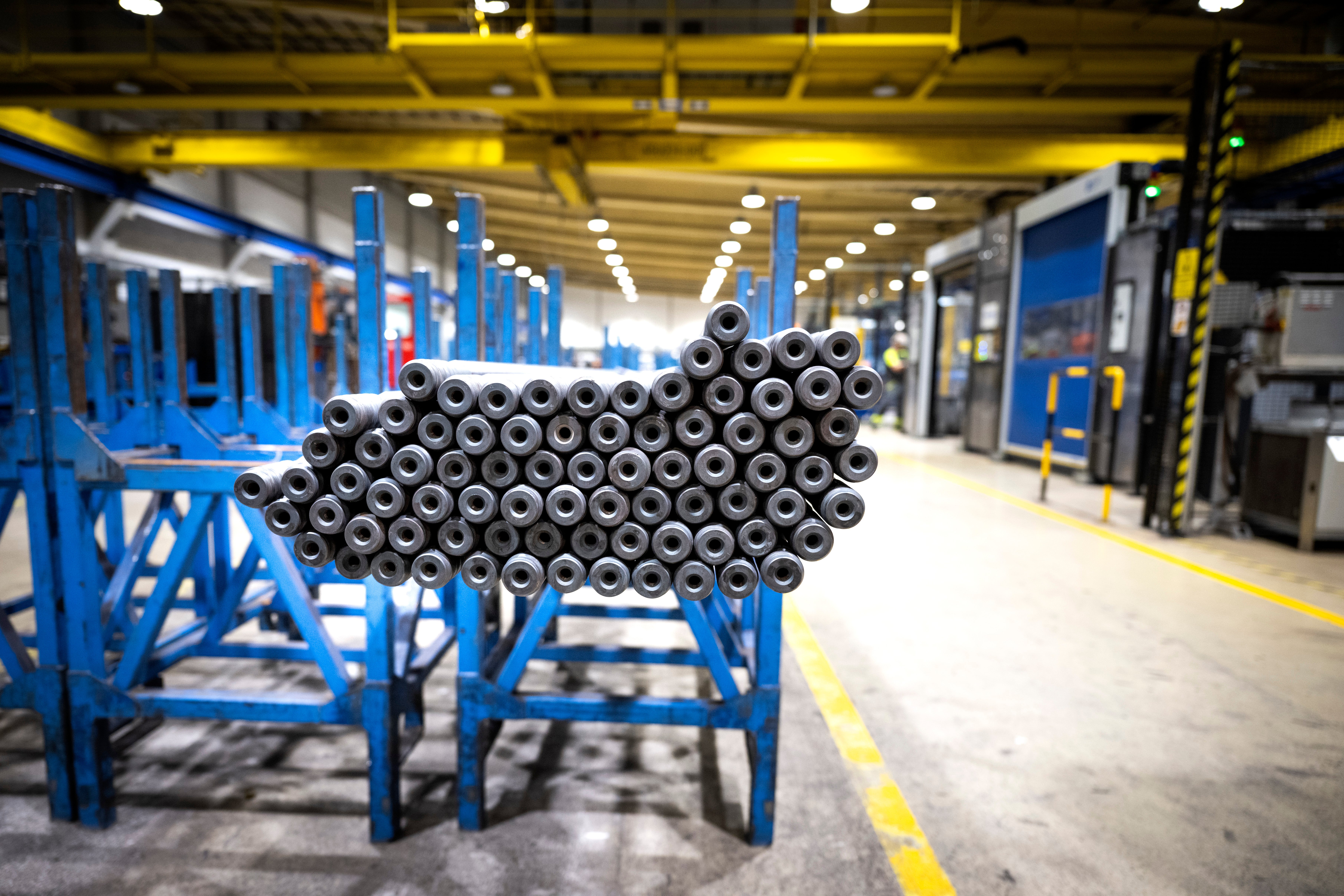 En stor bunt med metalliska rör arrangerade i en industriell fabrik med blå lagringssystem och gul markering på golvet.