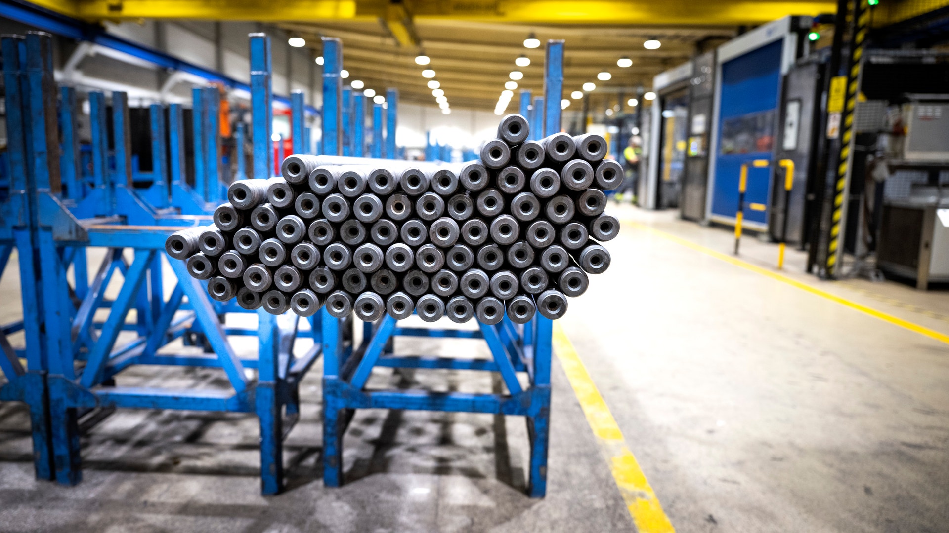En stor bunt med metalliska rör arrangerade i en industriell fabrik med blå lagringssystem och gul markering på golvet.