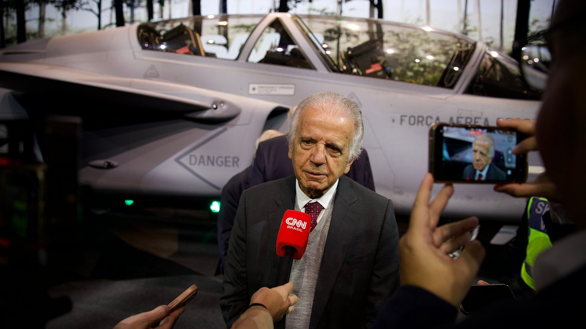 En äldre man i kostym intervjuas av journalister med mikrofoner. Ett militärflygplan syns i bakgrunden.