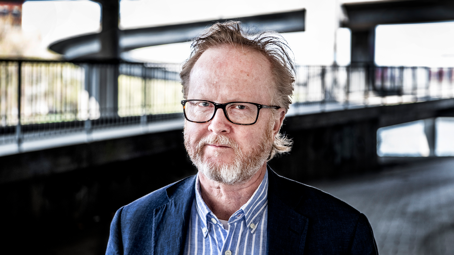 Porträttfoto av en man med glasögon och skägg, klädd i mörkblå kavaj och randig skjorta.