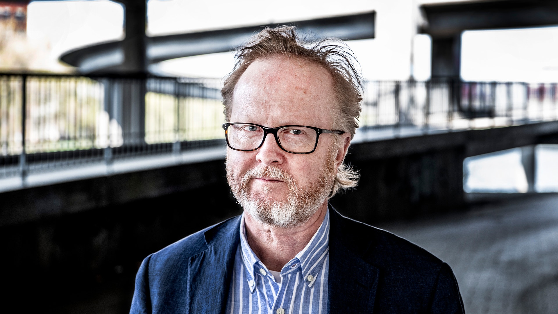 Porträttfoto av en man med glasögon och skägg, klädd i mörkblå kavaj och randig skjorta.