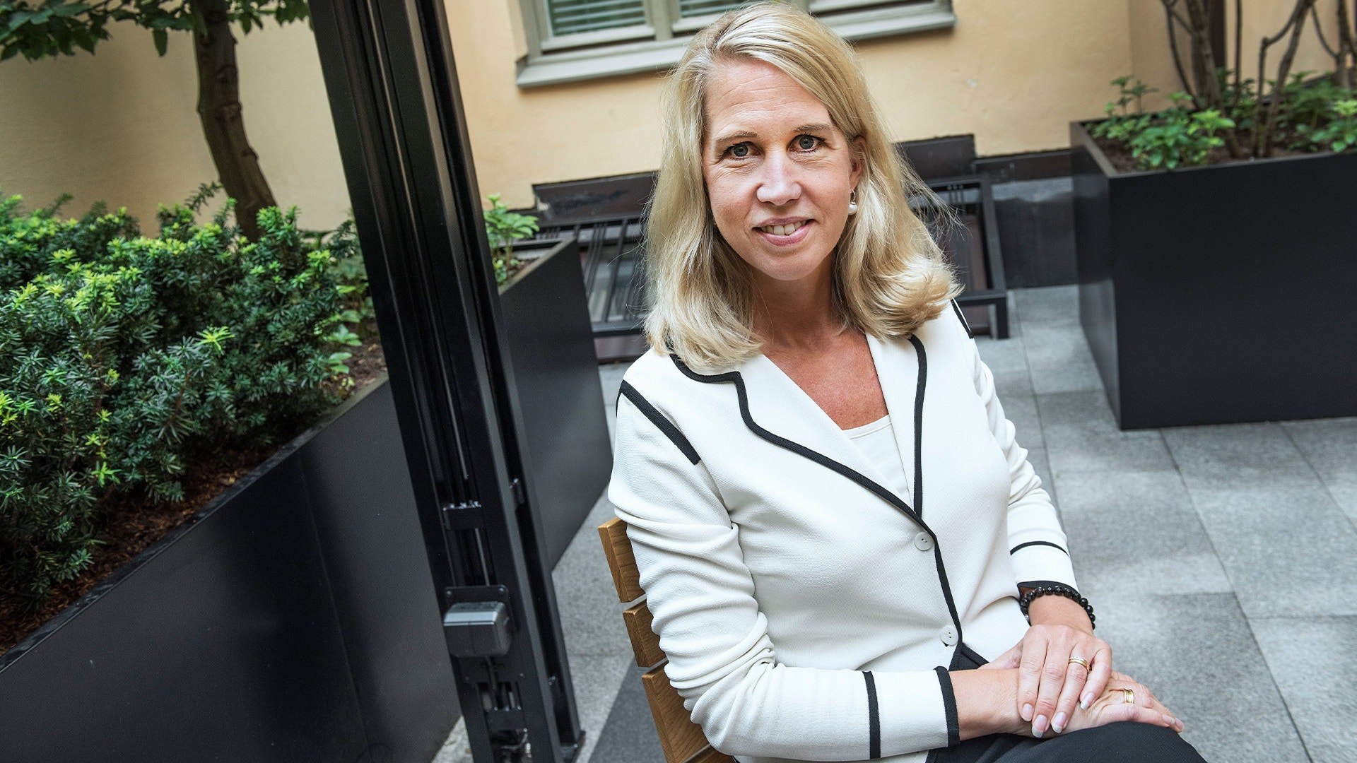 Porträtt av en kvinna som sitter på en bänk utomhus. Hon bär en vit blazer med svarta detaljer och tittar mot kameran. I bakgrunden syns gröna växter och en terass.