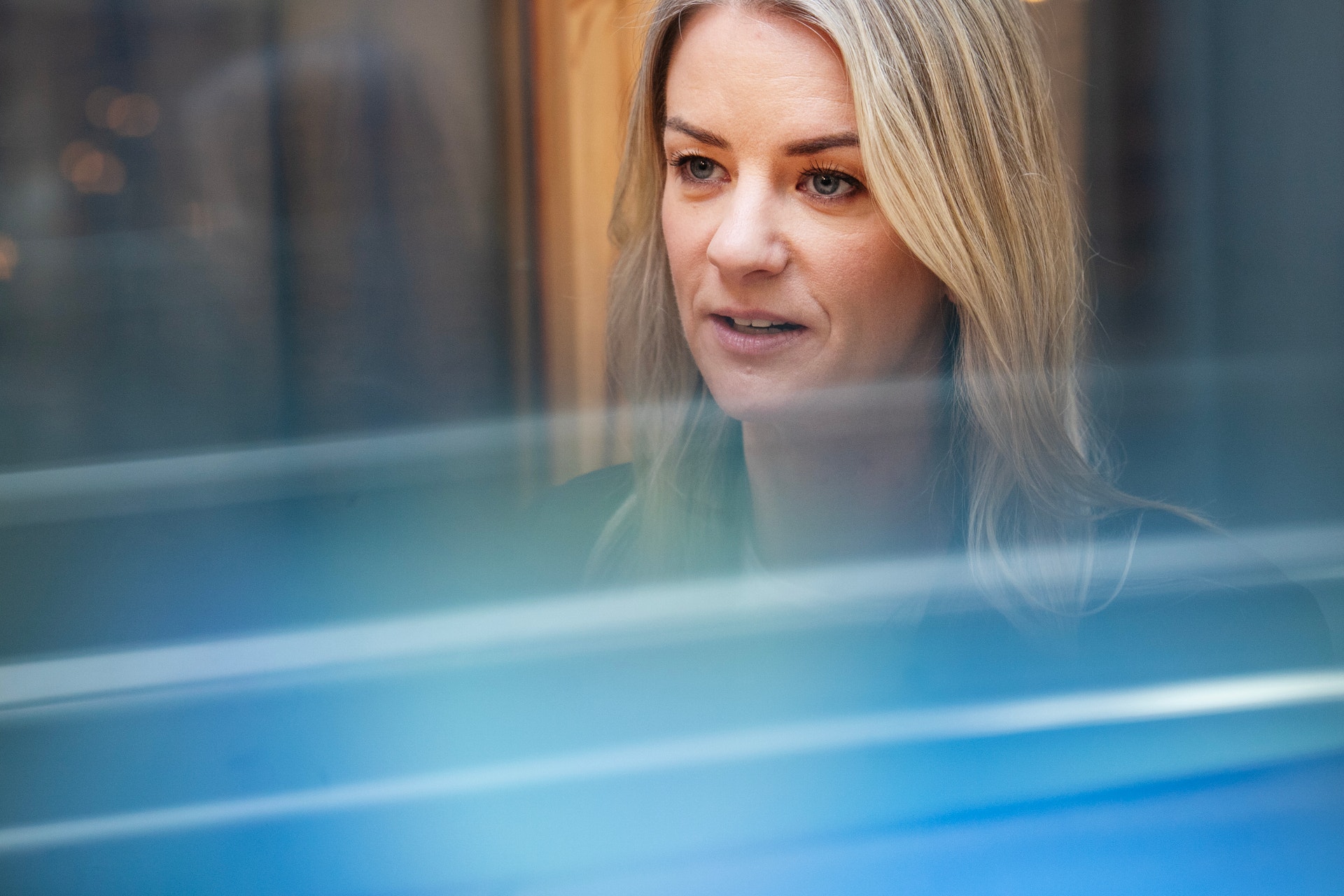 Porträtt av en kvinna med långt blont hår som tittar genom ett glasfönster med blå reflexer.
