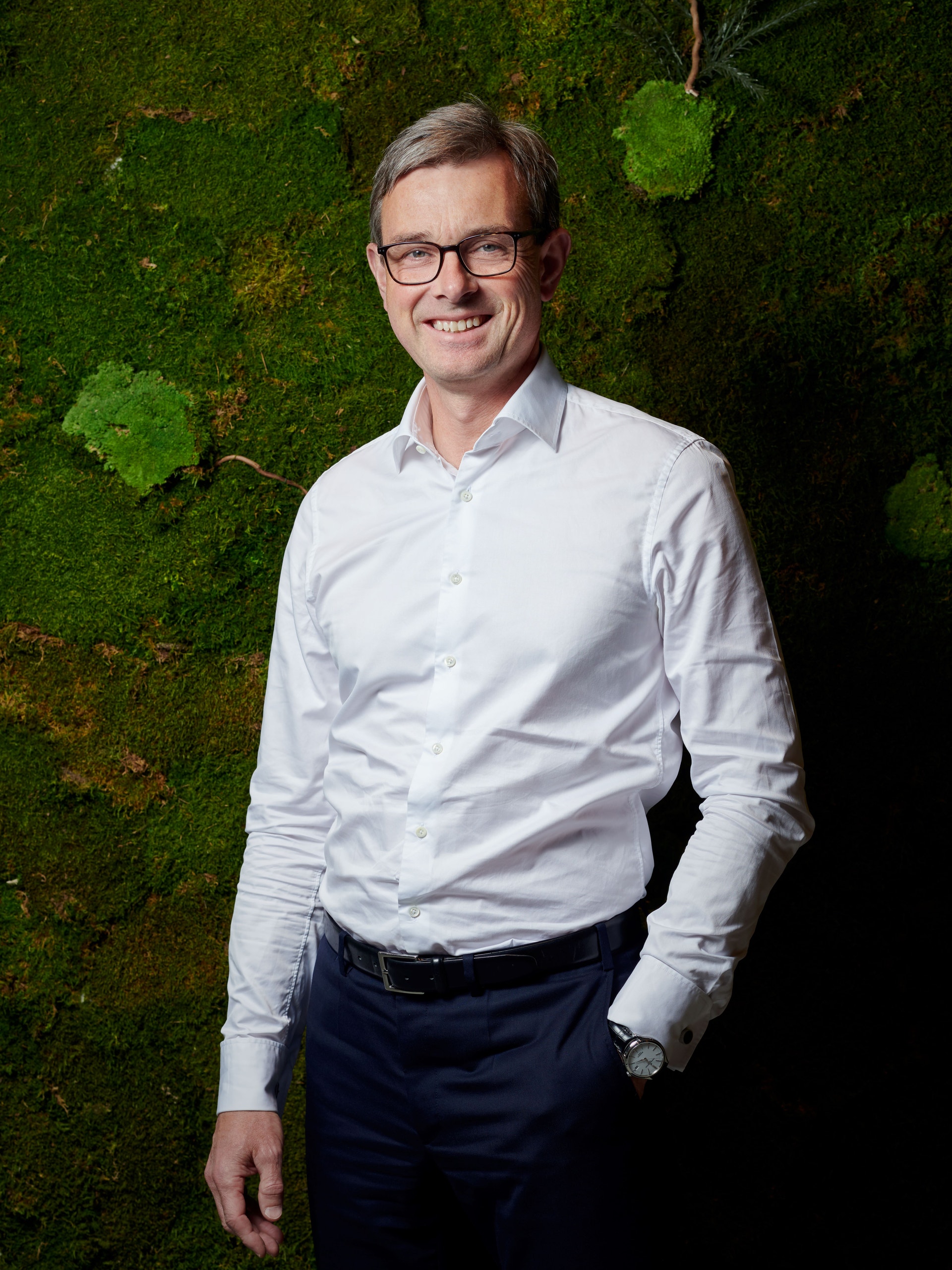 Porträtt av en man med glasögon i vit skjorta framför en vägg med mossa och växter.