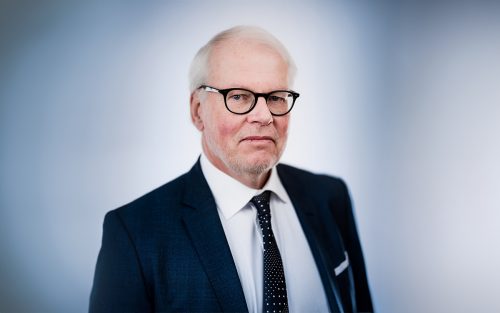 Porträtt av en äldre man med glasögon, vit skjorta och mörkblå kostym med slips.